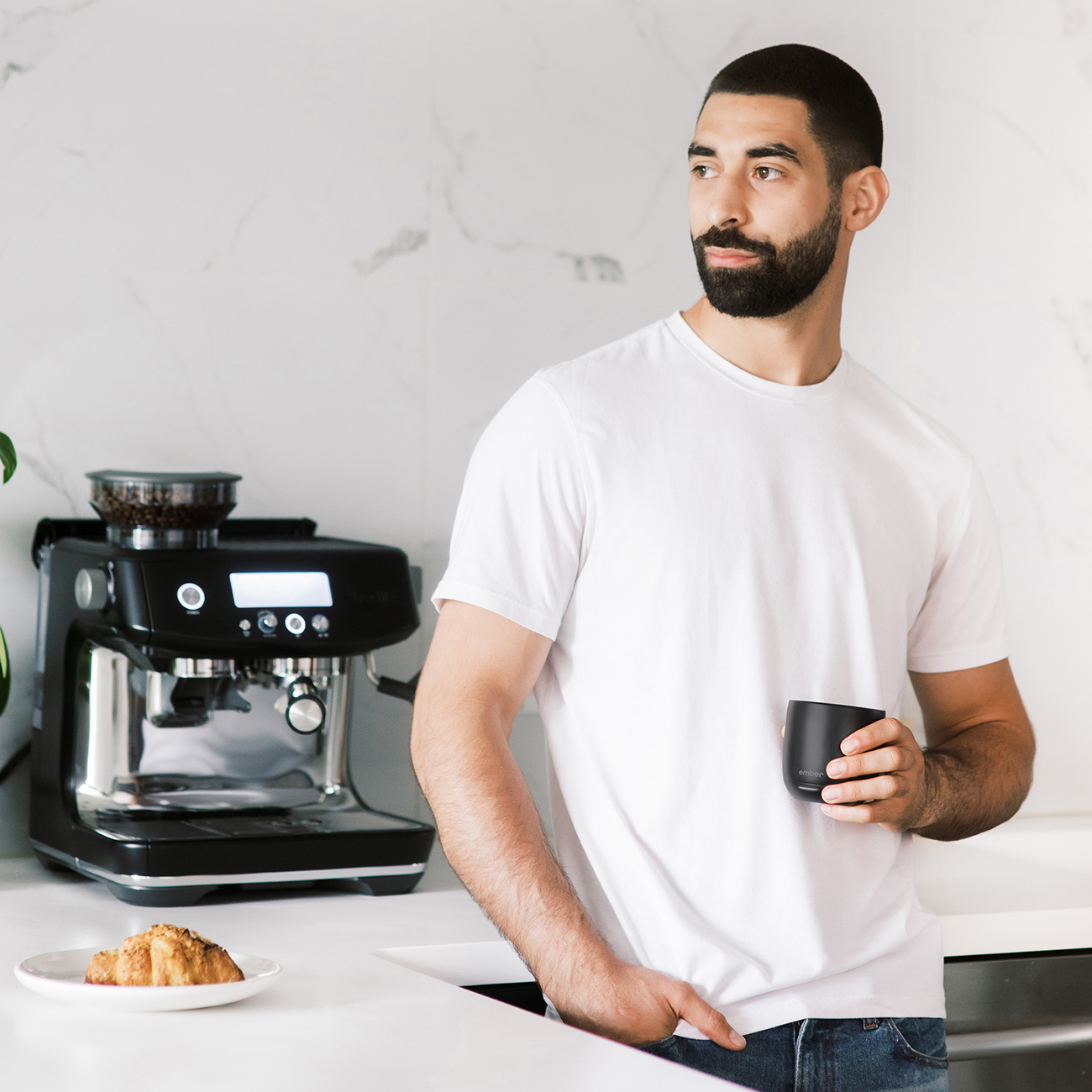 Uomo con la barba che tiene una tazza nera, vicino a una macchina per caffè espresso su un piano di lavoro bianco.