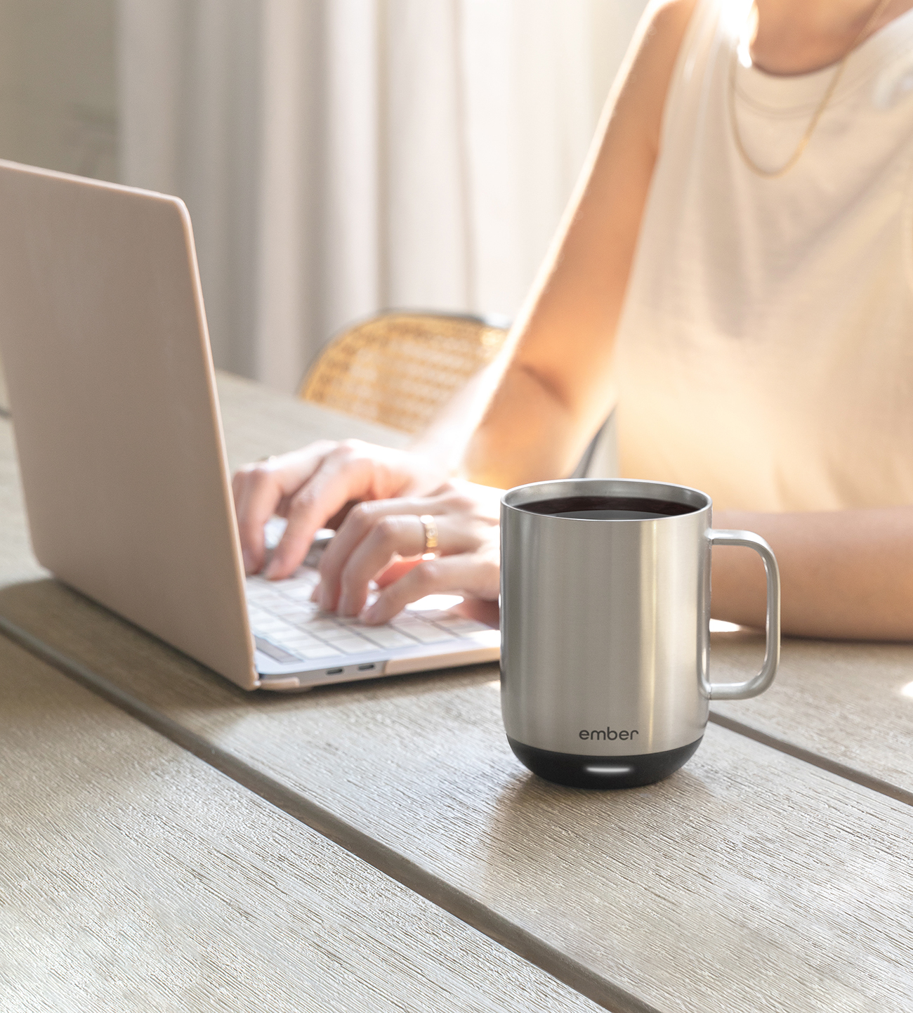 Una persona usando una laptop con una taza plateada con base negra sobre una mesa de madera. Luz cálida.