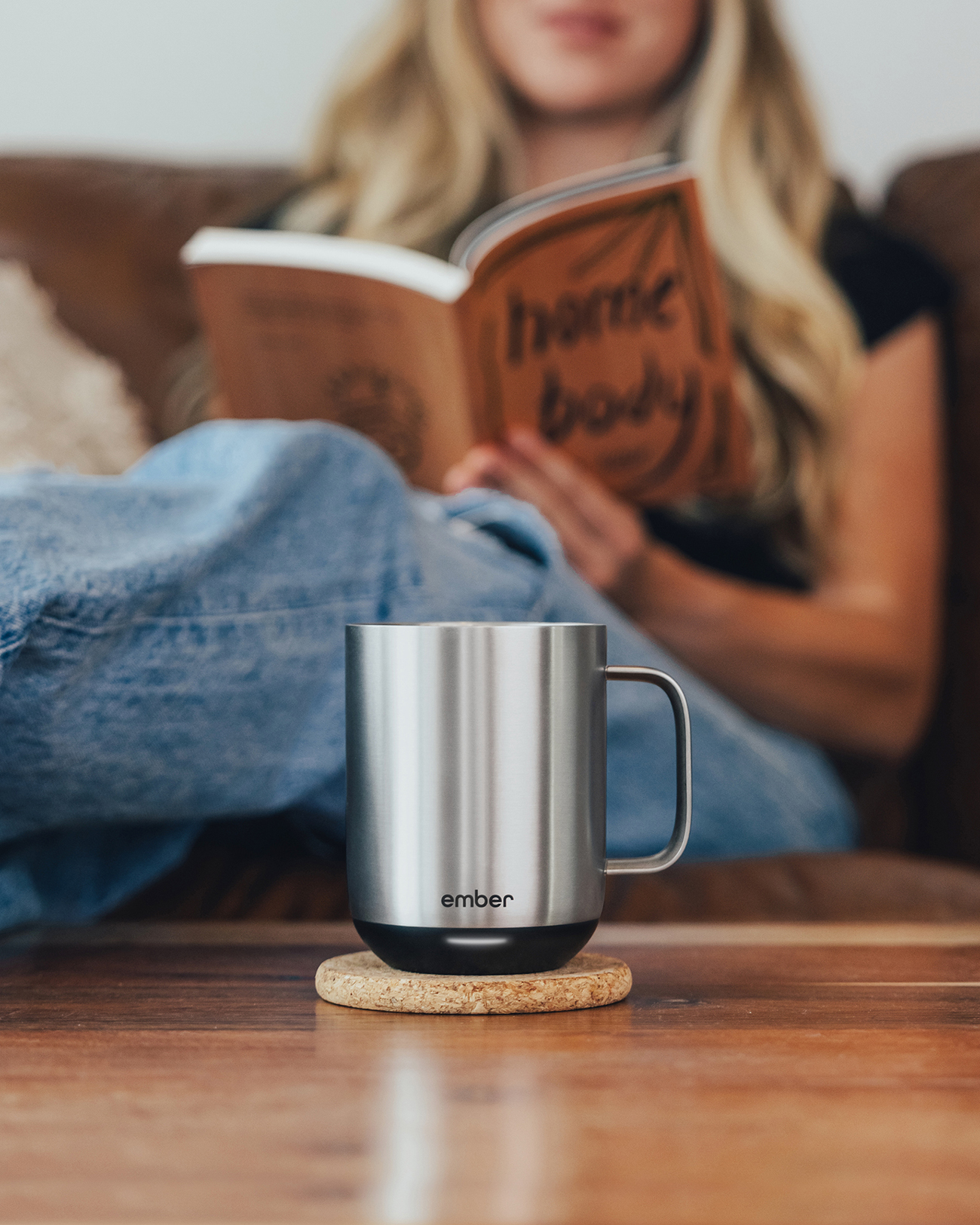 Una taza de acero inoxidable sobre un posavasos. Una mujer lee un libro en el fondo, sentada en un sofá.