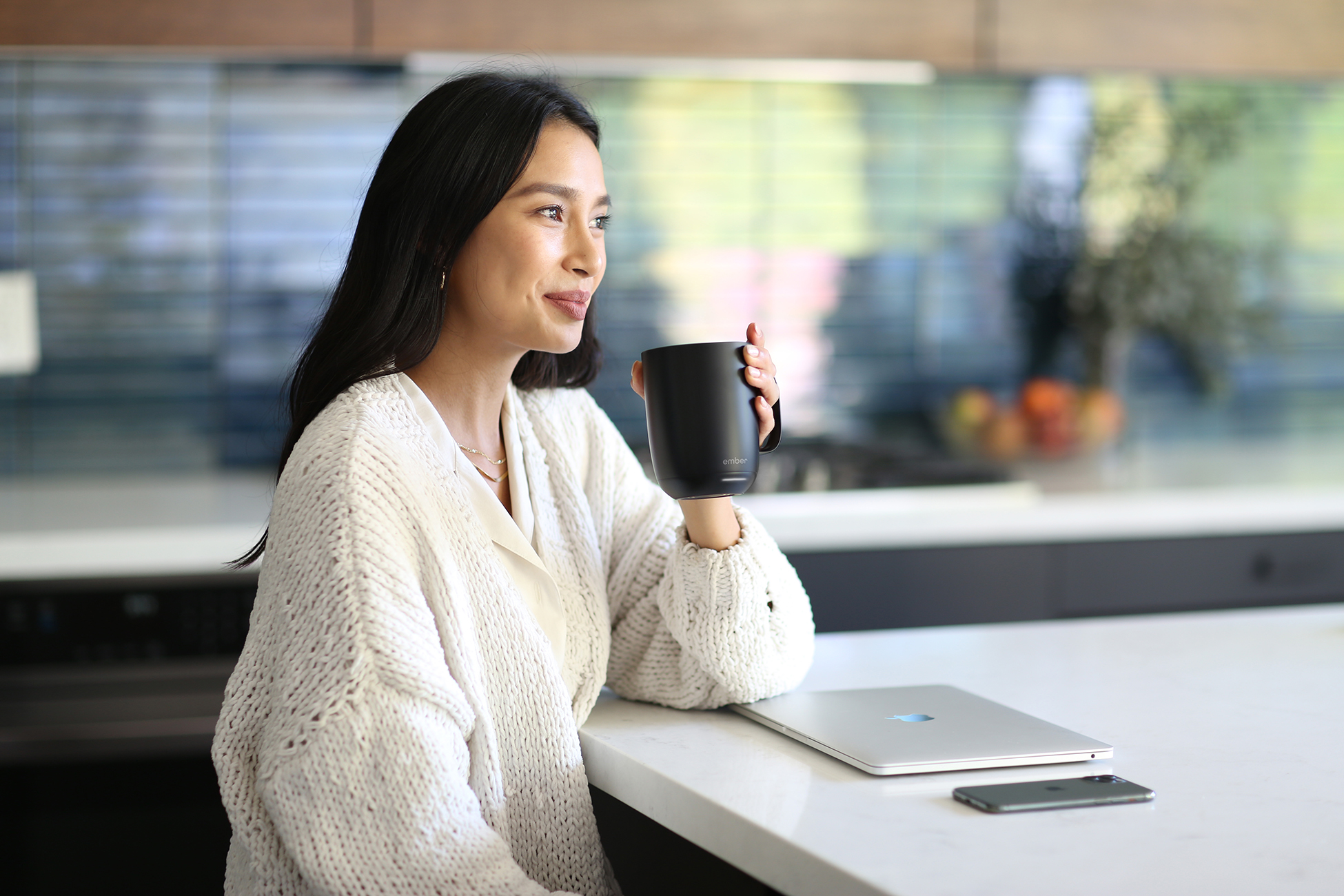 Una donna che tiene una tazza nera, con un laptop e un telefono su un bancone in una cucina moderna.