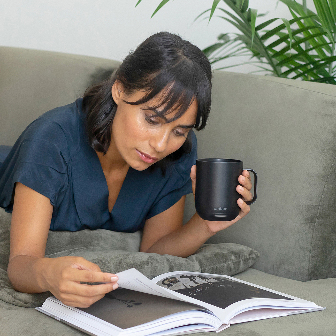 Una donna che legge un libro, tenendo una tazza nera, su un divano grigio.