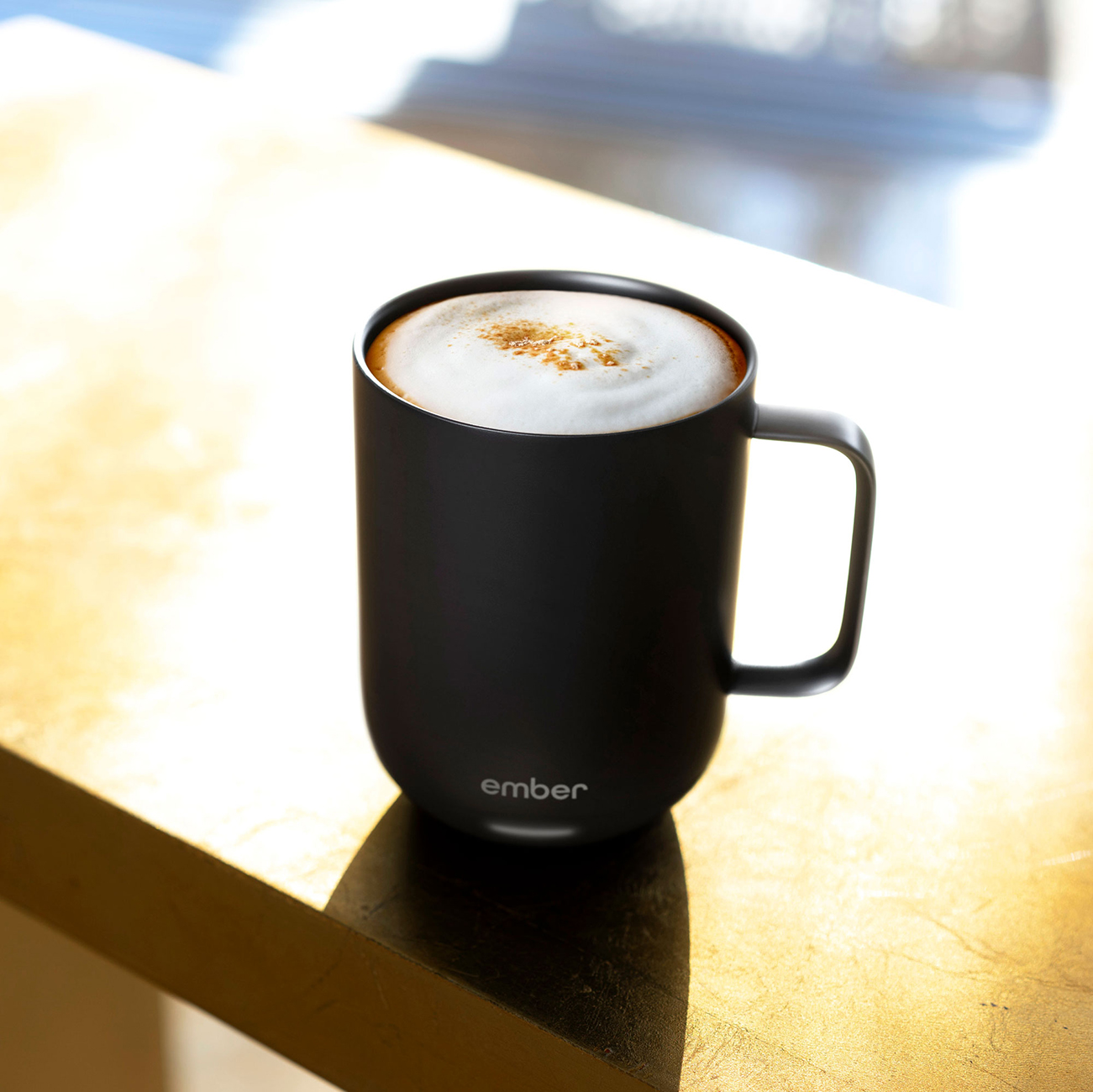Tazza nera con caffè e schiuma su superficie dorata. Luce intensa dalla finestra.