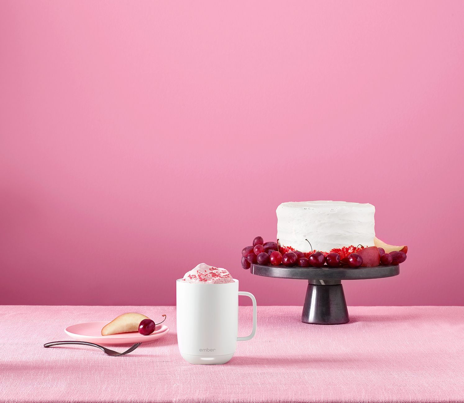 Uno sfondo rosa con una torta bianca su un supporto, una tazza con panna montata, frutta e un piatto con una pera.