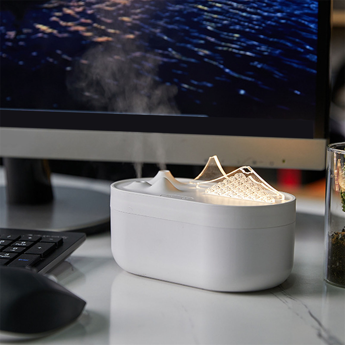 Weißer Luftbefeuchter auf einem Schreibtisch mit bergförmigem Design, der Dampf abgibt. Im Hintergrund befinden sich ein Monitor und eine Tastatur.
