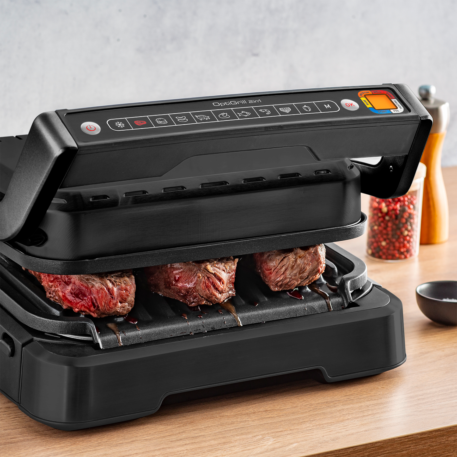 Czarny grill z stekami w środku. Panel sterowania widoczny na górze, drewniana powierzchnia poniżej. Urządzenie kuchenne.