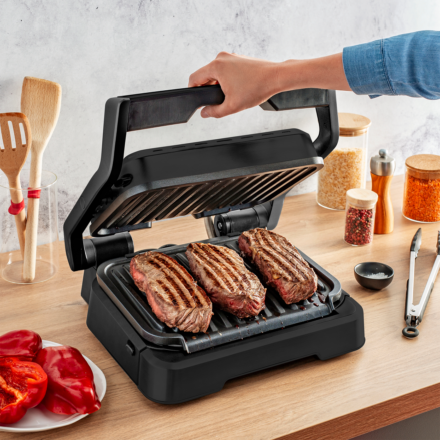 Czarny grill z trzema grillowanymi stekami. Ręka podnosi pokrywę. Otoczenie kuchni z drewnianą powierzchnią.