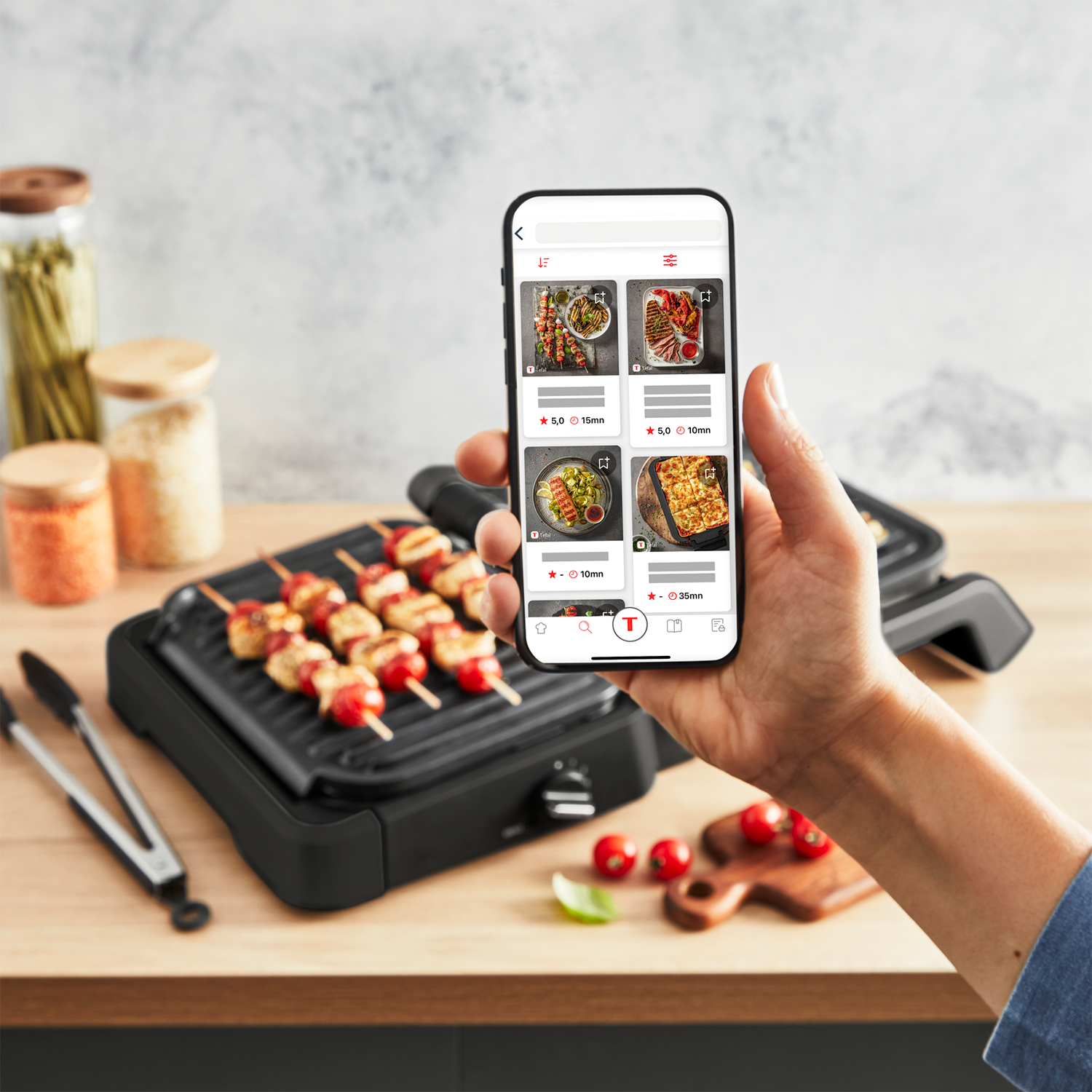 Ręka trzymająca telefon z przepisami na grilla. Grill i szaszłyki z jedzeniem w tle. Otoczenie kuchni.