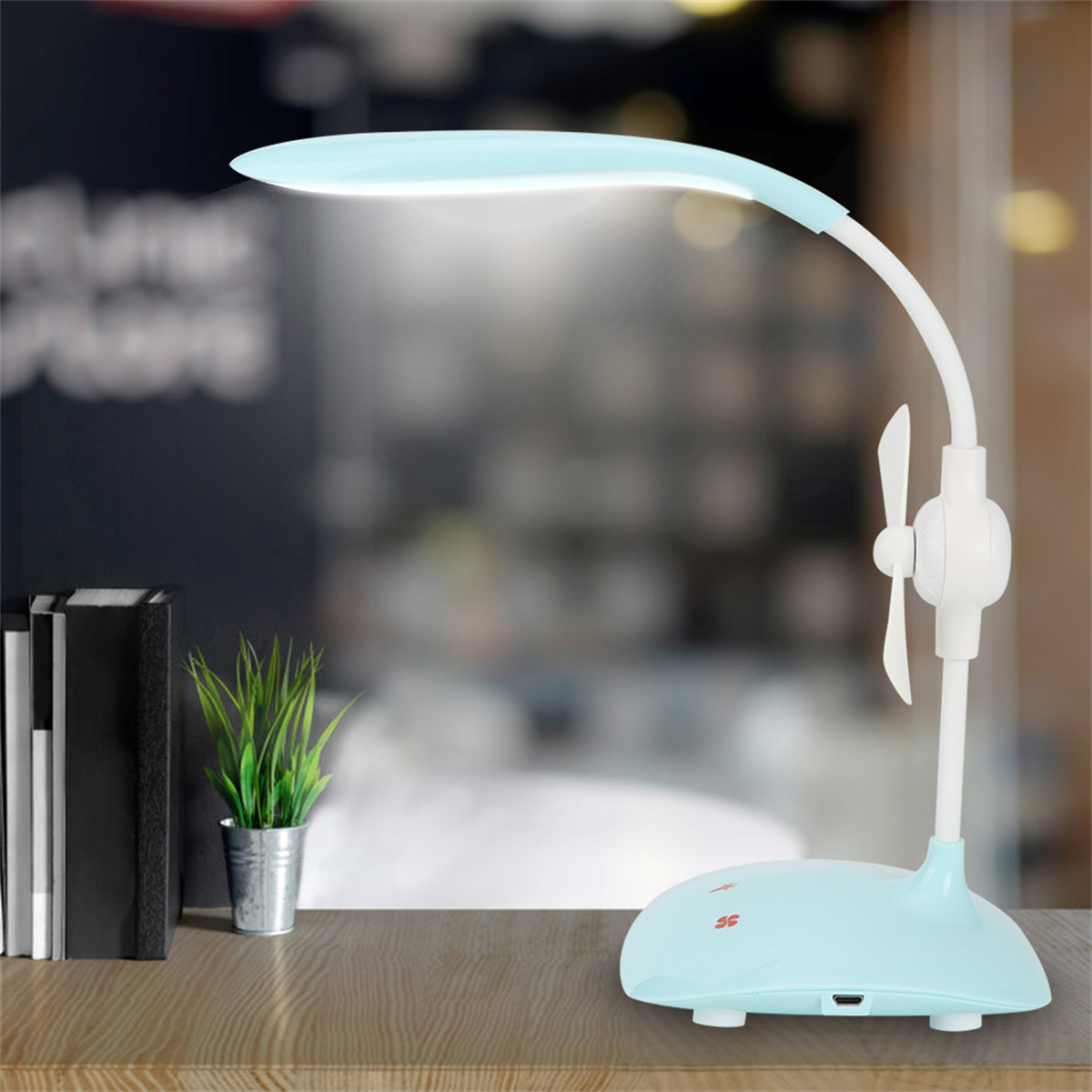 Eine hellblaue Schreibtischlampe mit Ventilator, neben Büchern und einer kleinen Pflanze.