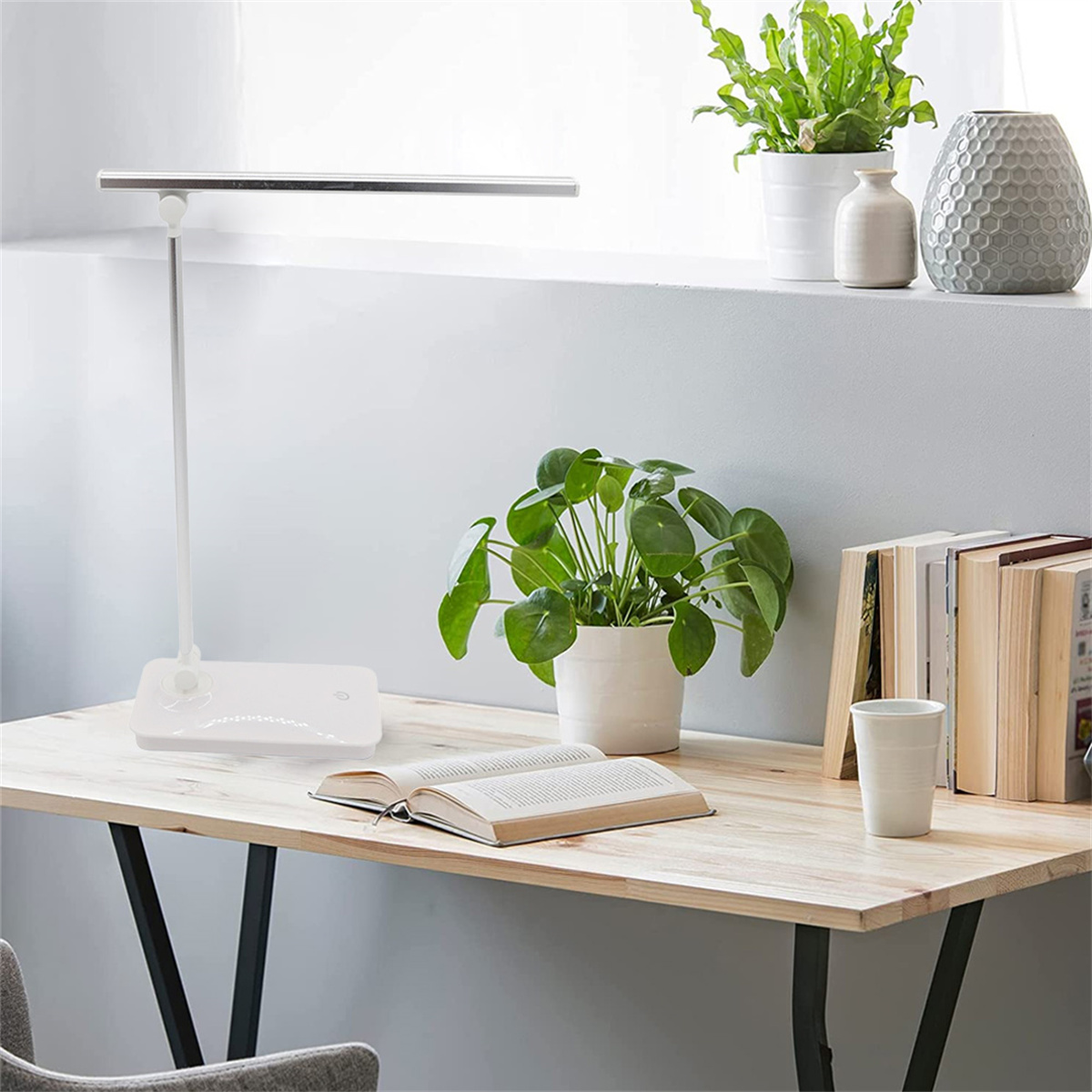 Eine Schreibtischlampe mit weißem Sockel und einem langen rechteckigen Licht. Bücher, Pflanzen und eine Tasse auf dem Schreibtisch.