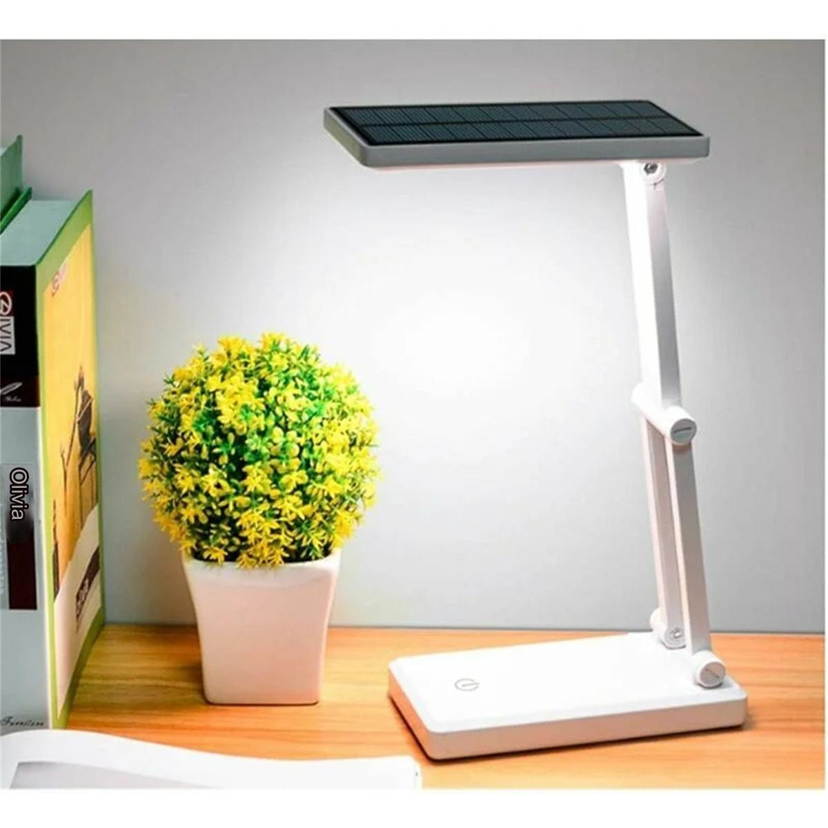 Eine weiße Schreibtischlampe mit Solarpanel oben. Auf dem Schreibtisch stehen eine Pflanze und Bücher.