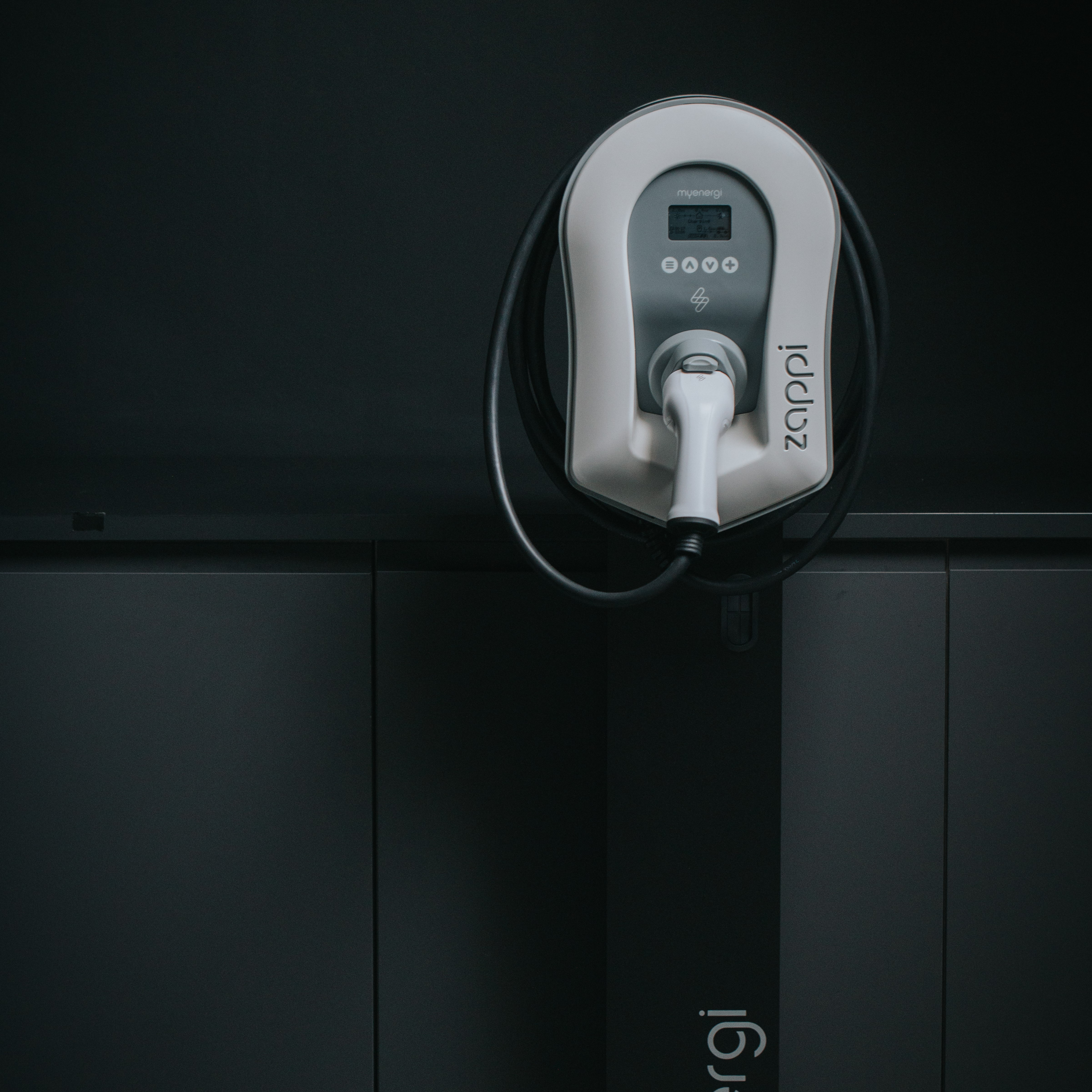 White and gray EV charger mounted on a dark wall, with a black charging cable.