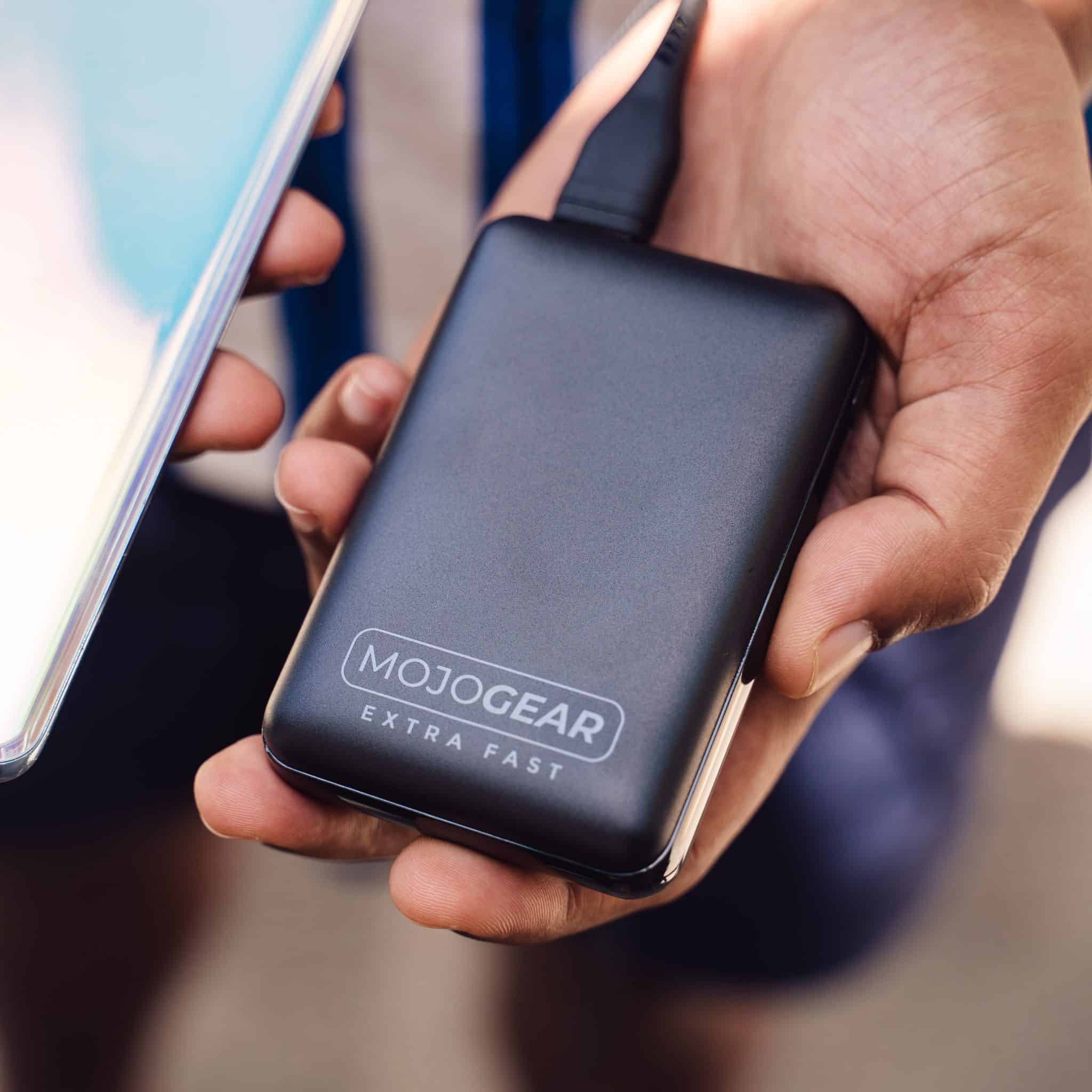 Chargeur portable noir tenu par une main à côté d'un téléphone. Le chargeur indique MOJOGEAR EXTRA FAST.