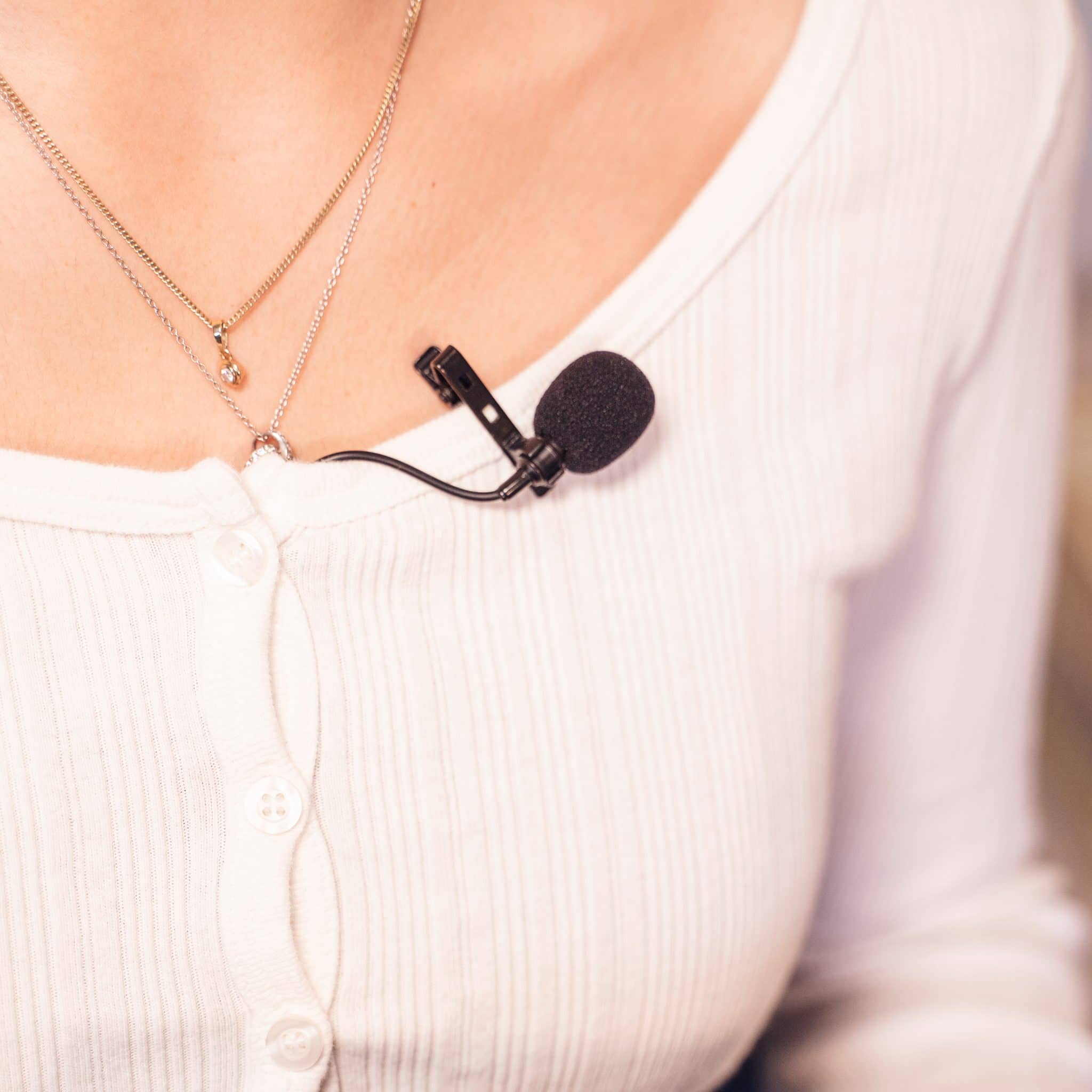 Borst van een vrouw met een dasspeldmicrofoon aan haar witte overhemd en ketting.
