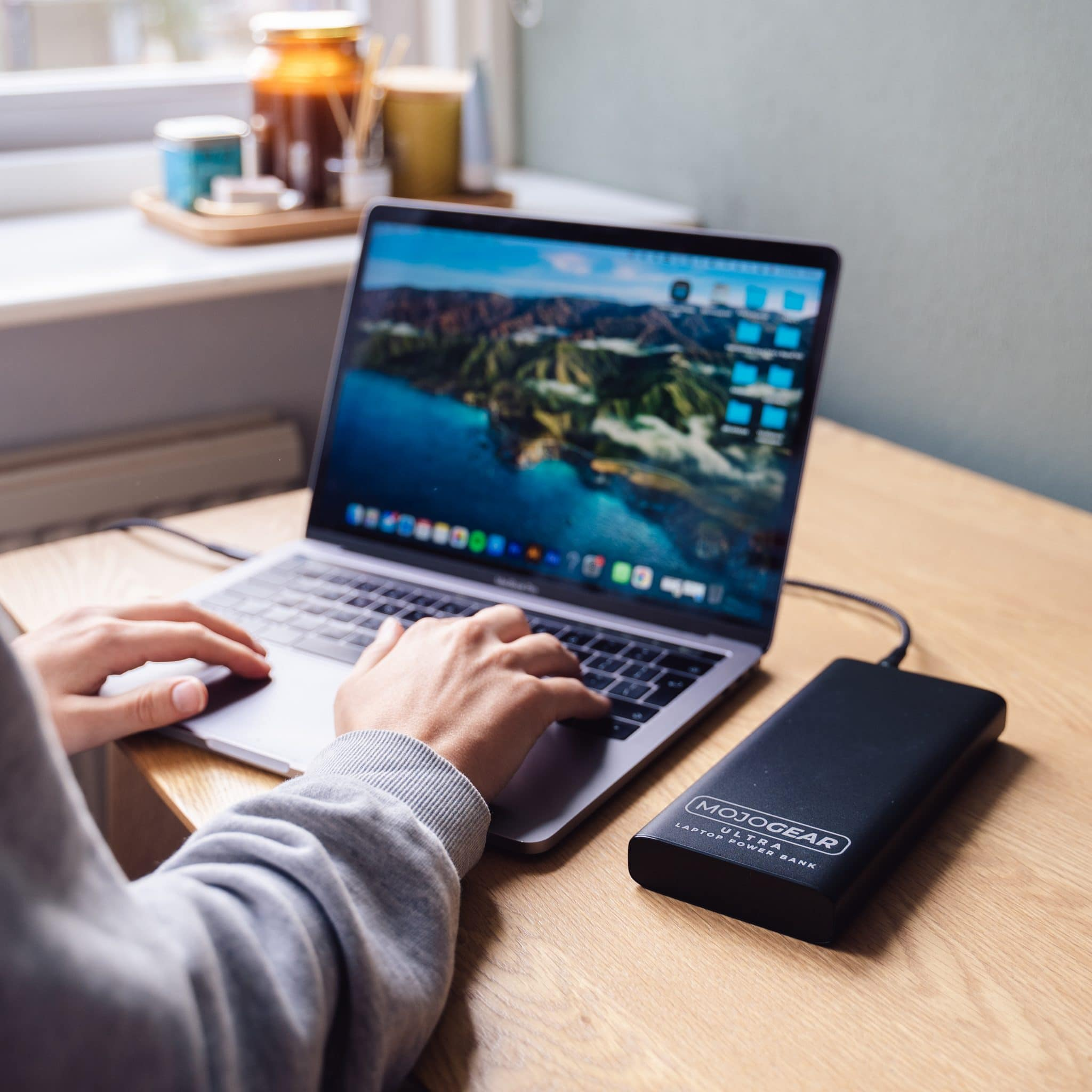 Een persoon die een laptop gebruikt. Een zwarte powerbank staat naast de laptop op een houten tafel.