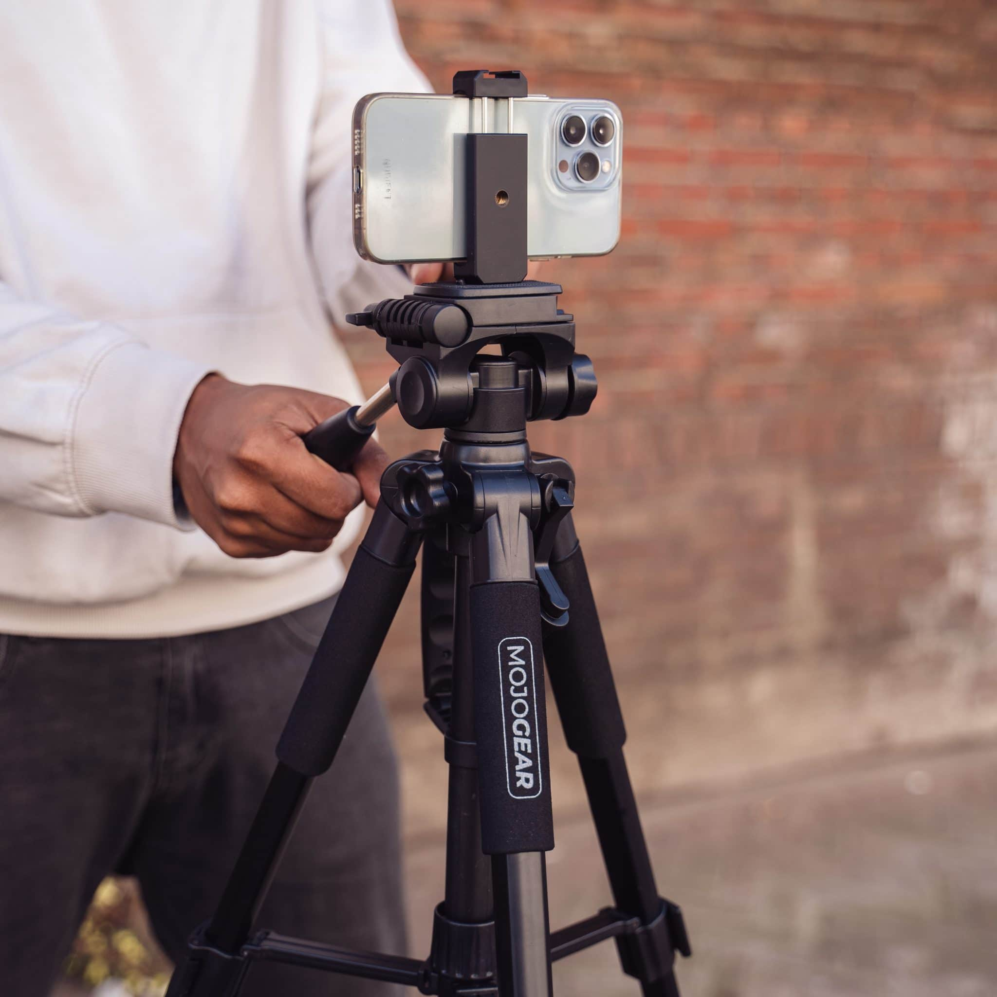A person holding a tripod with a phone attached, against a brick wall background.