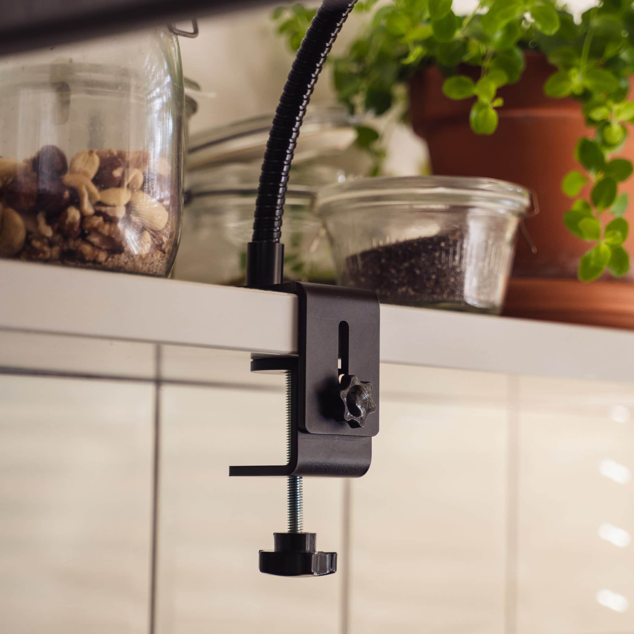 Een zwarte klem met een flexibele arm die een plank vasthoudt. Potten en planten zijn zichtbaar.