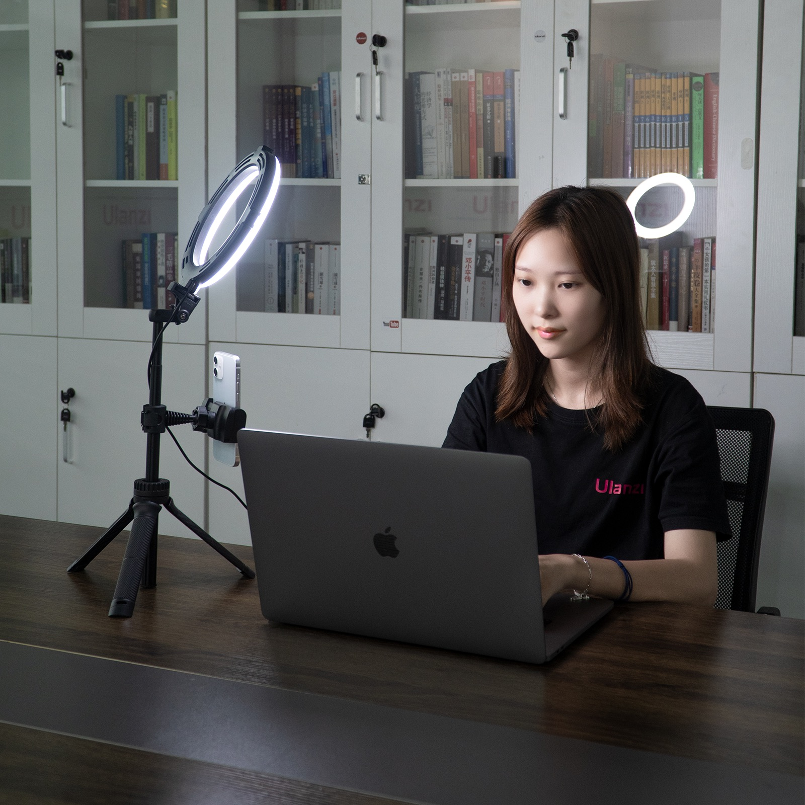 Woman working on a laptop with a ring light and phone holder set up. Bookshelf in the background.