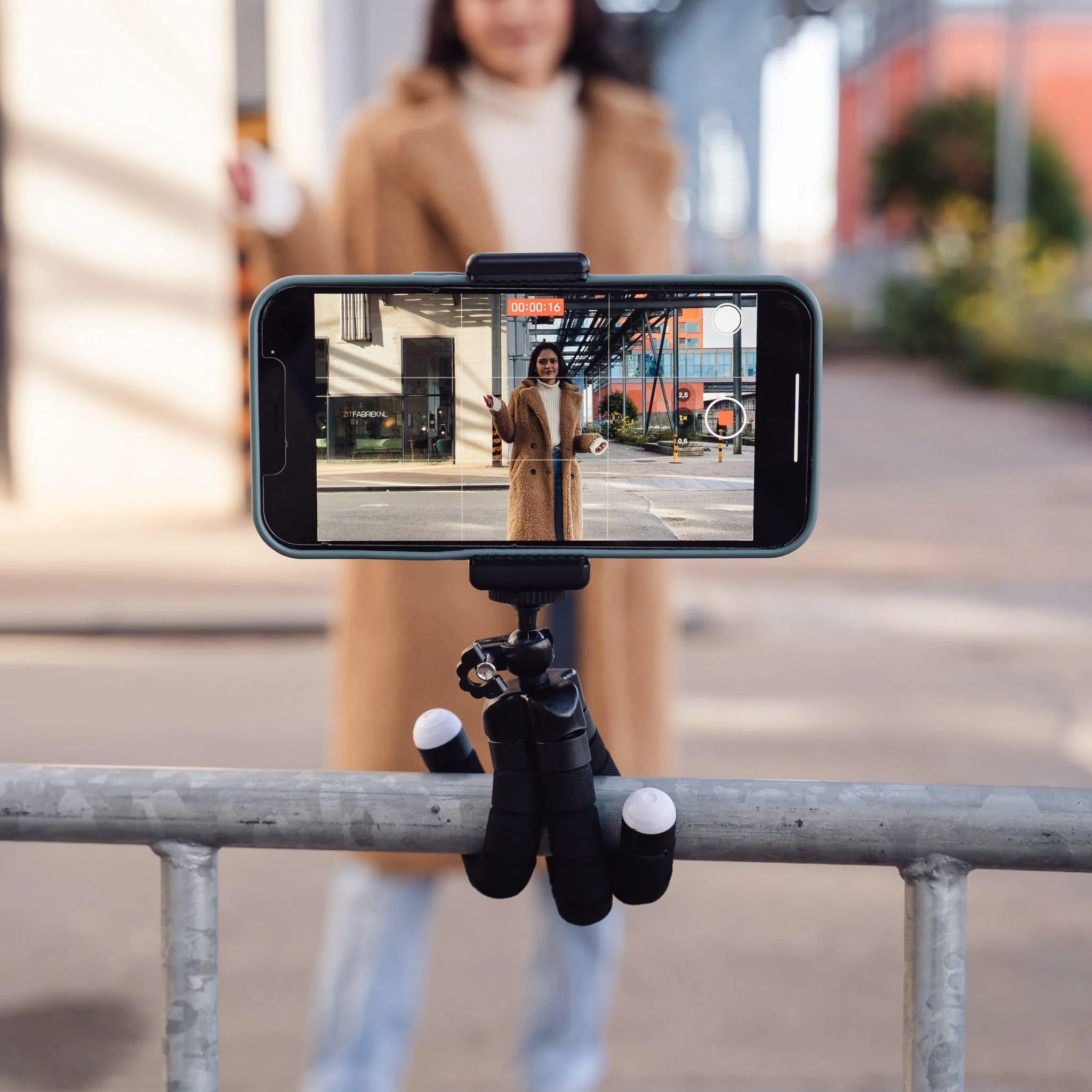 A phone on a tripod records a woman in a coat, outdoors.