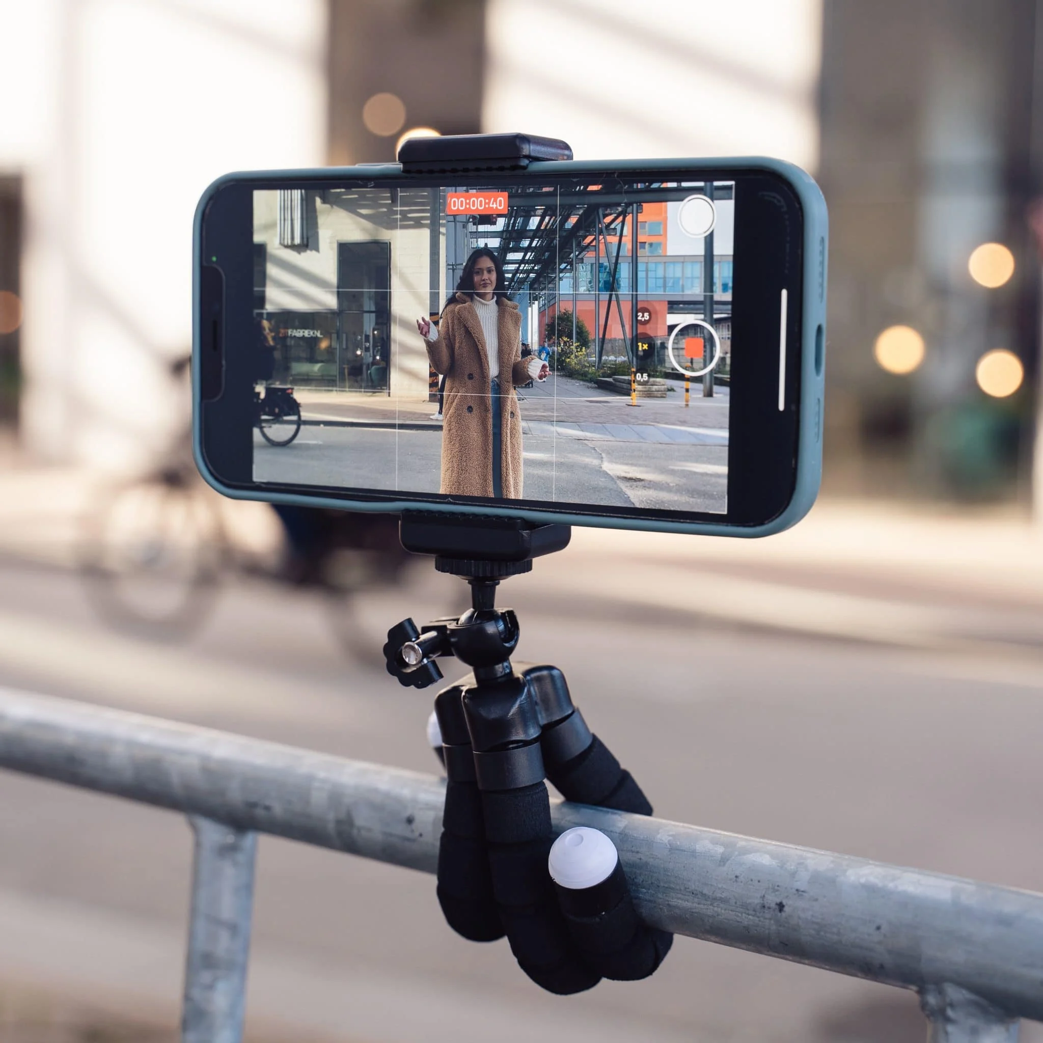 A phone is mounted on a tripod, displaying a video of a woman in a coat, filming on a street.