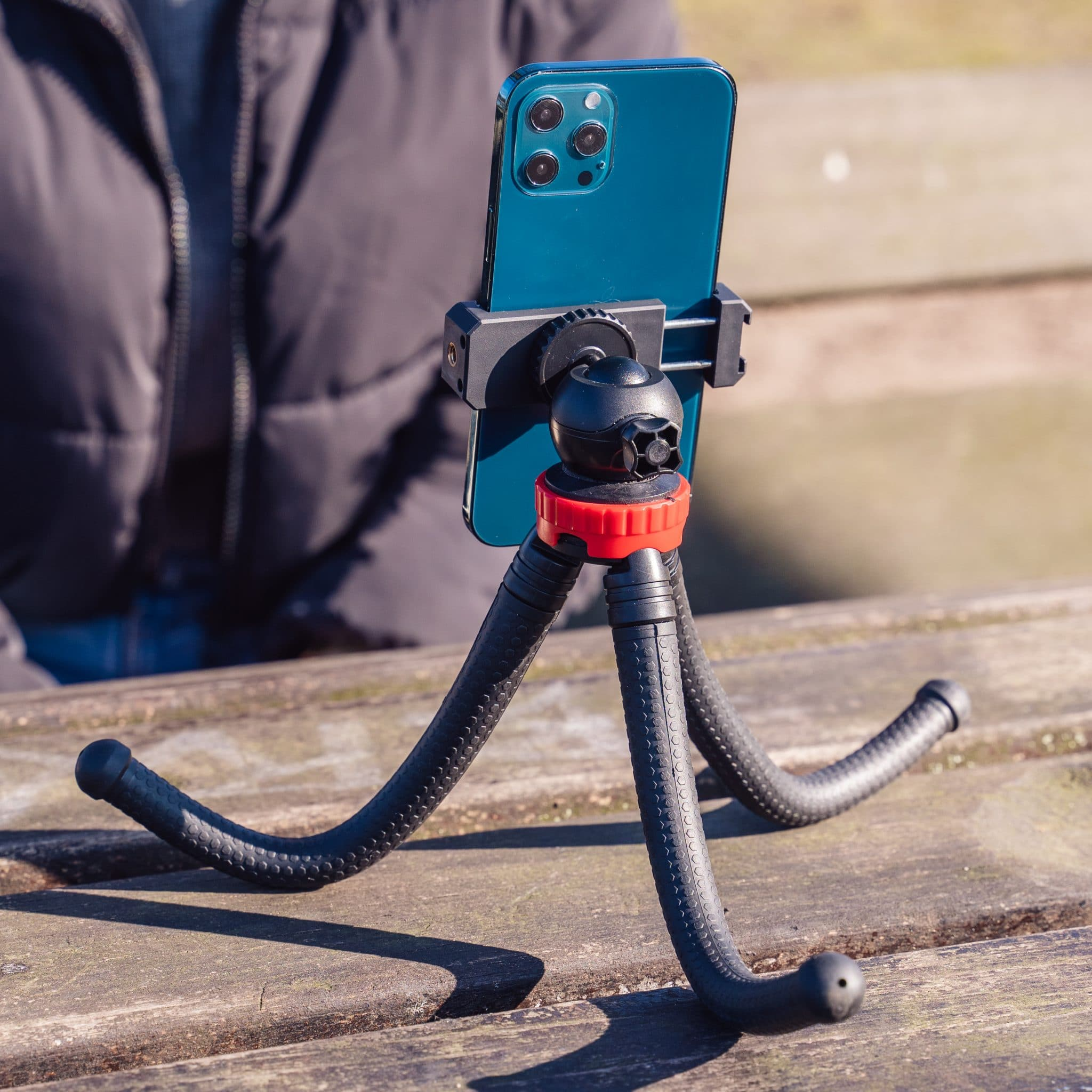Un téléphone bleu sarcelle sur un trépied. Le trépied a des pieds noirs flexibles et une bague de réglage rouge. Une personne est en arrière-plan.