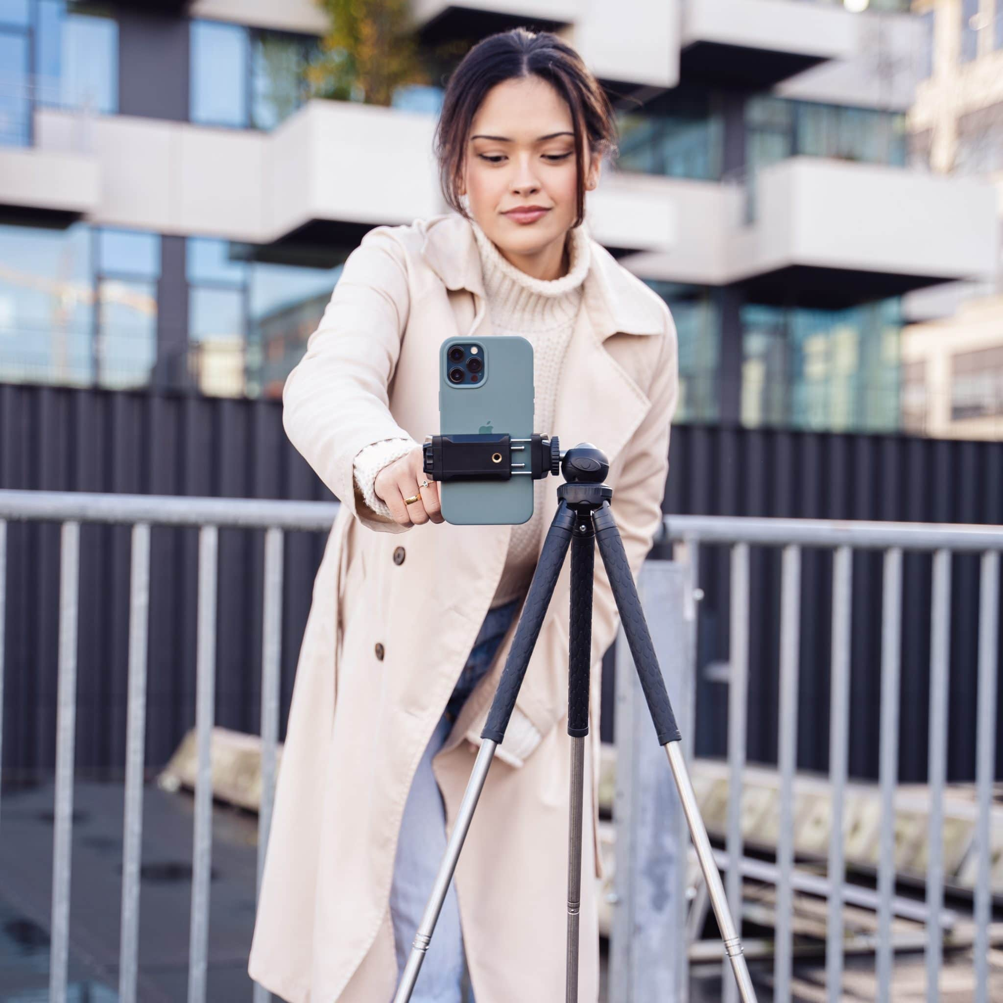 A woman in a beige coat sets up a phone on a tripod outdoors, near a metal fence.