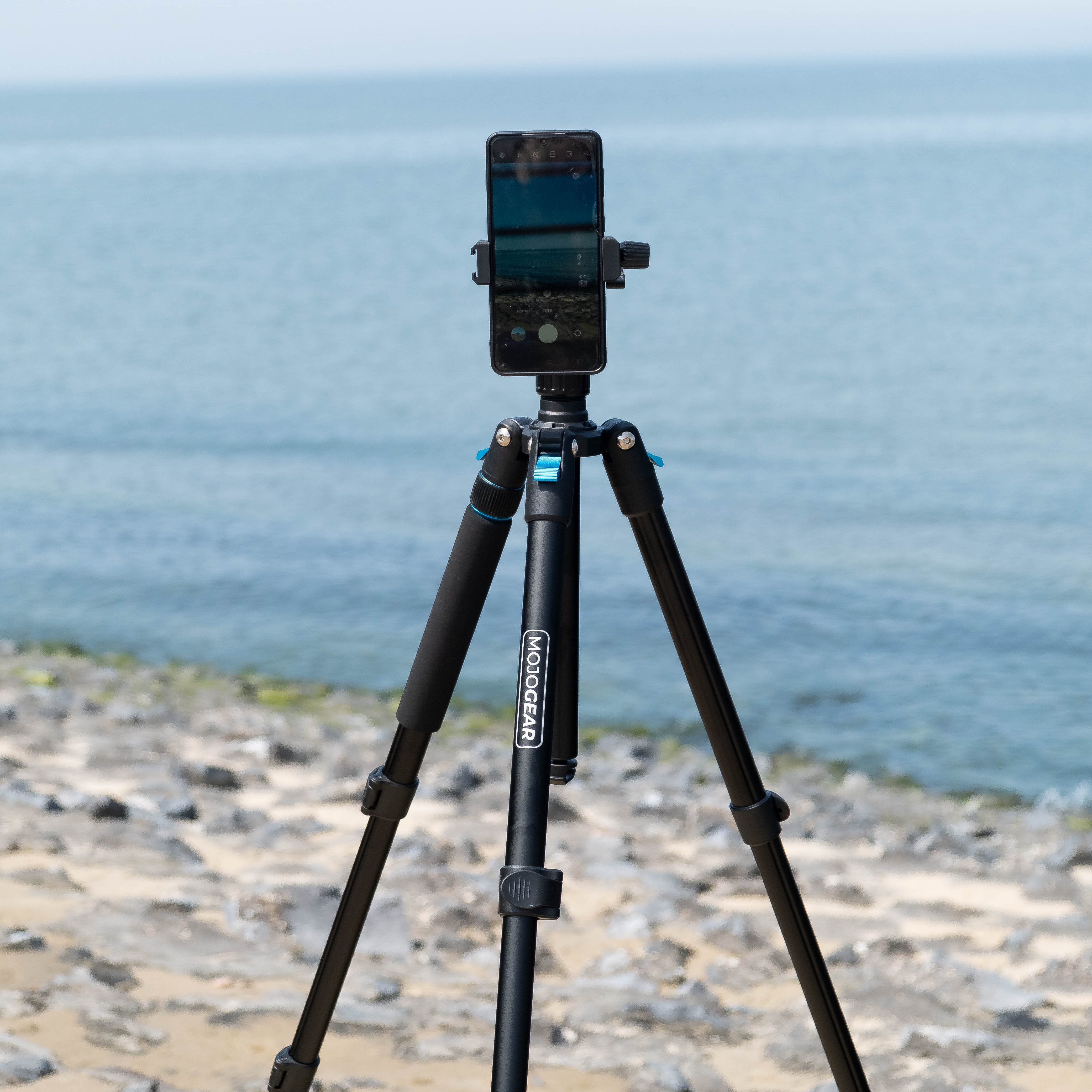 A black tripod holds a phone, capturing a beach and ocean view. The sky is clear blue.