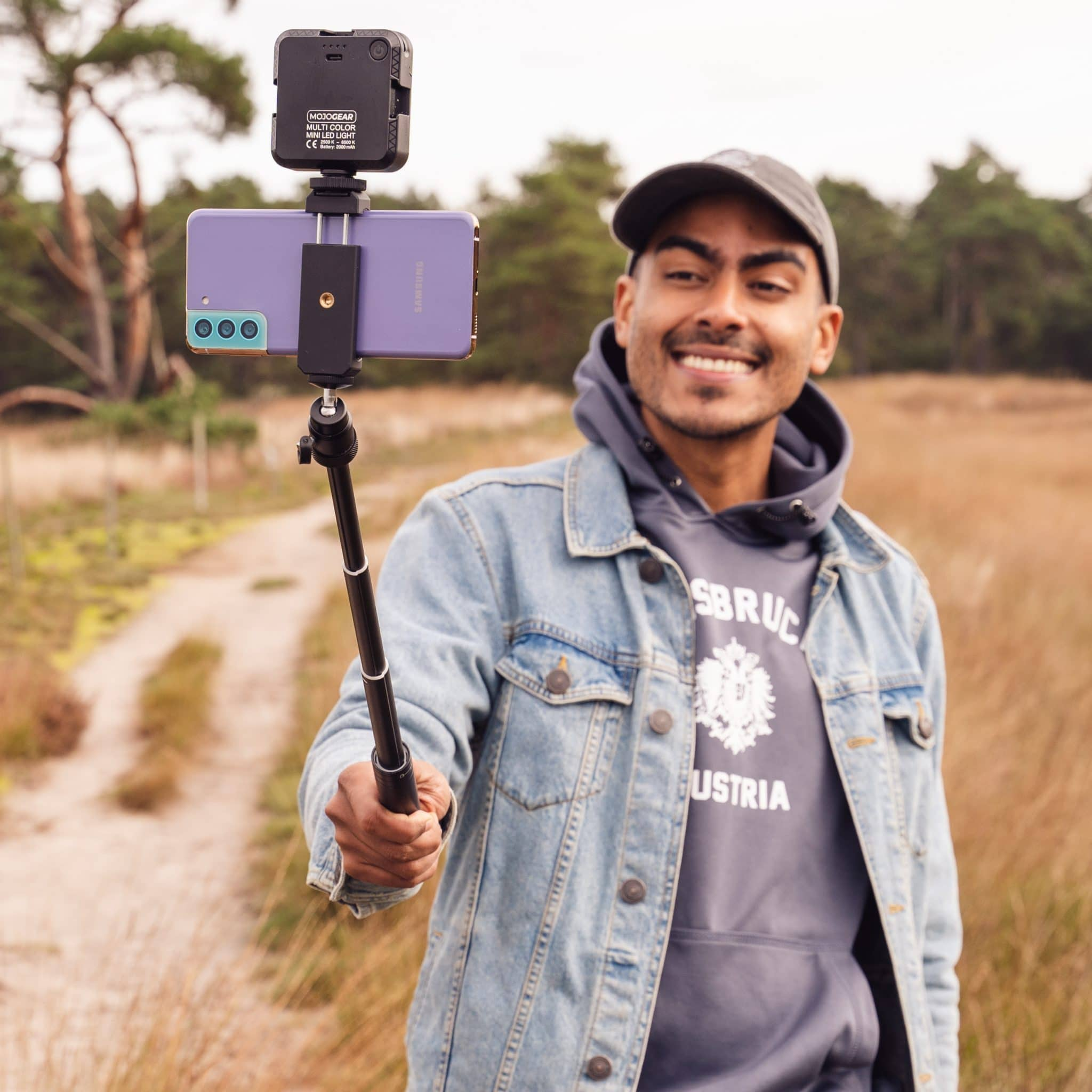 Mann hält Telefon auf Selfie-Stick. Feldhintergrund mit Bäumen. Lächelt.