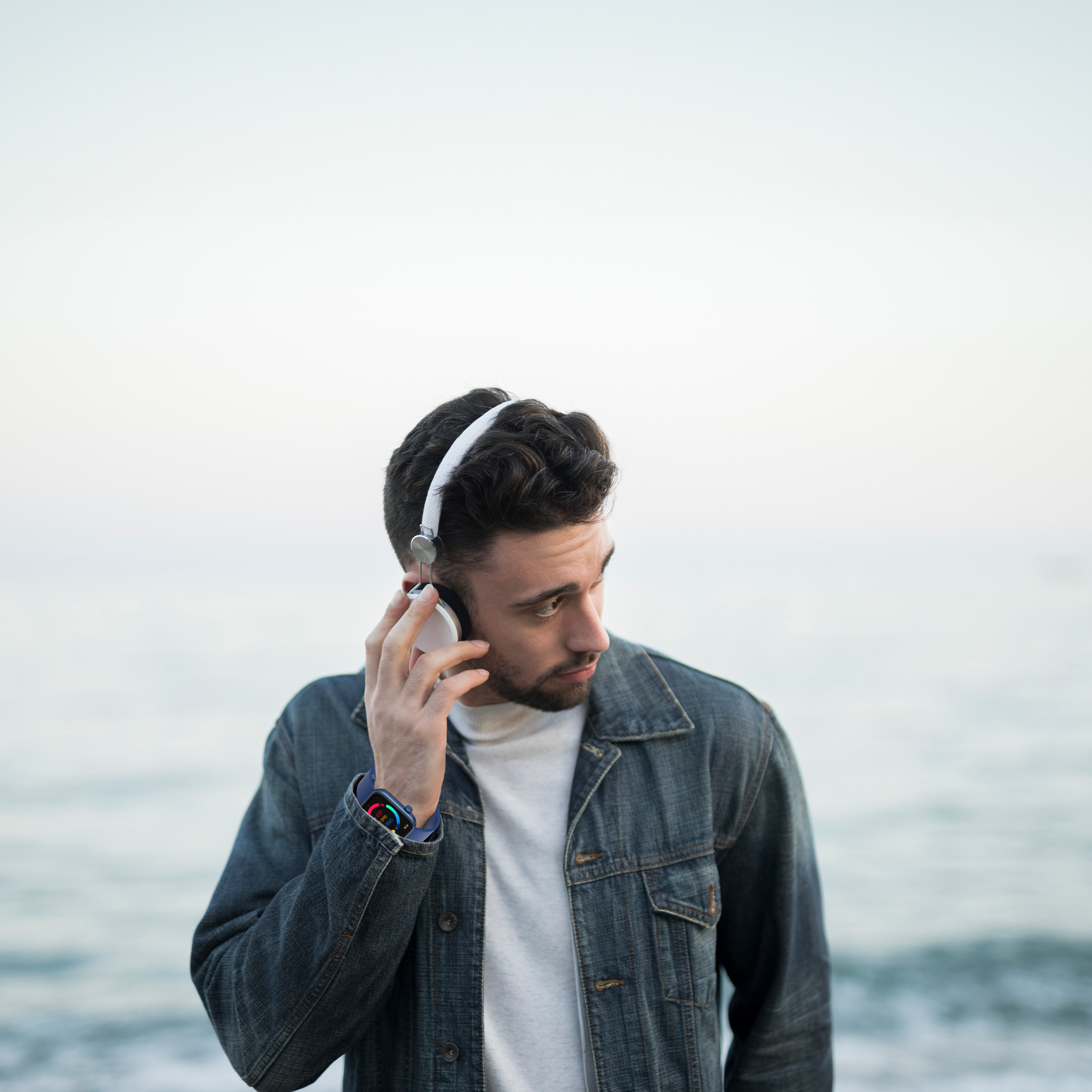 Mann mit Jeansjacke und Kopfhörern, der am Meer zur Seite blickt.