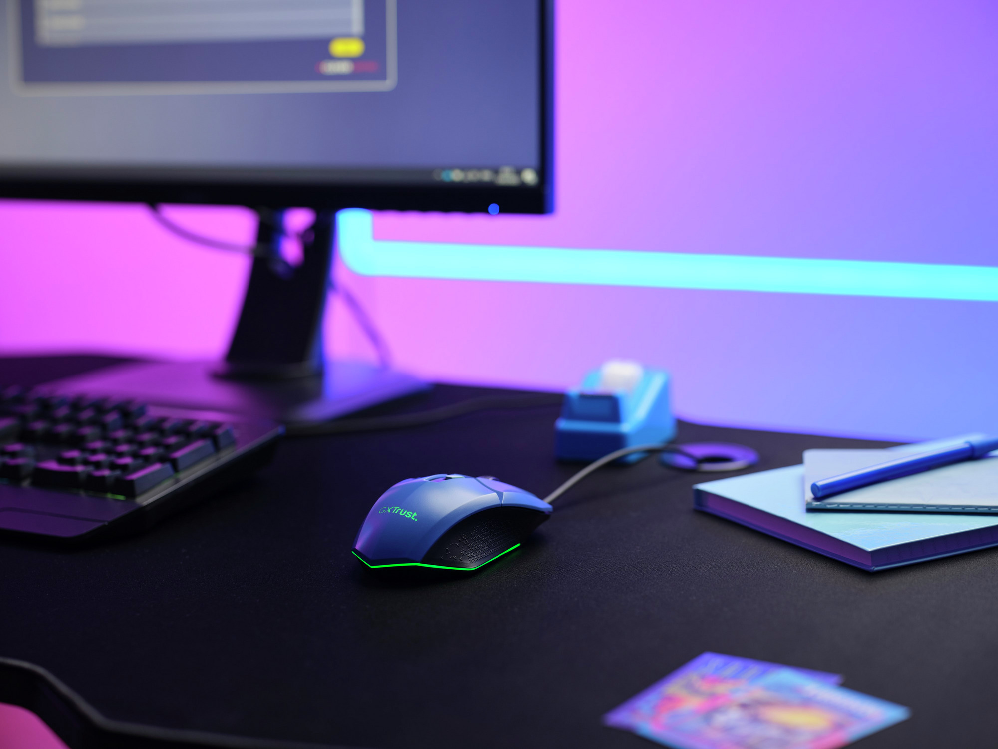 Niebieska mysz komputerowa na czarnym biurku. Inne obiekty: klawiatura, monitor i notatnik.