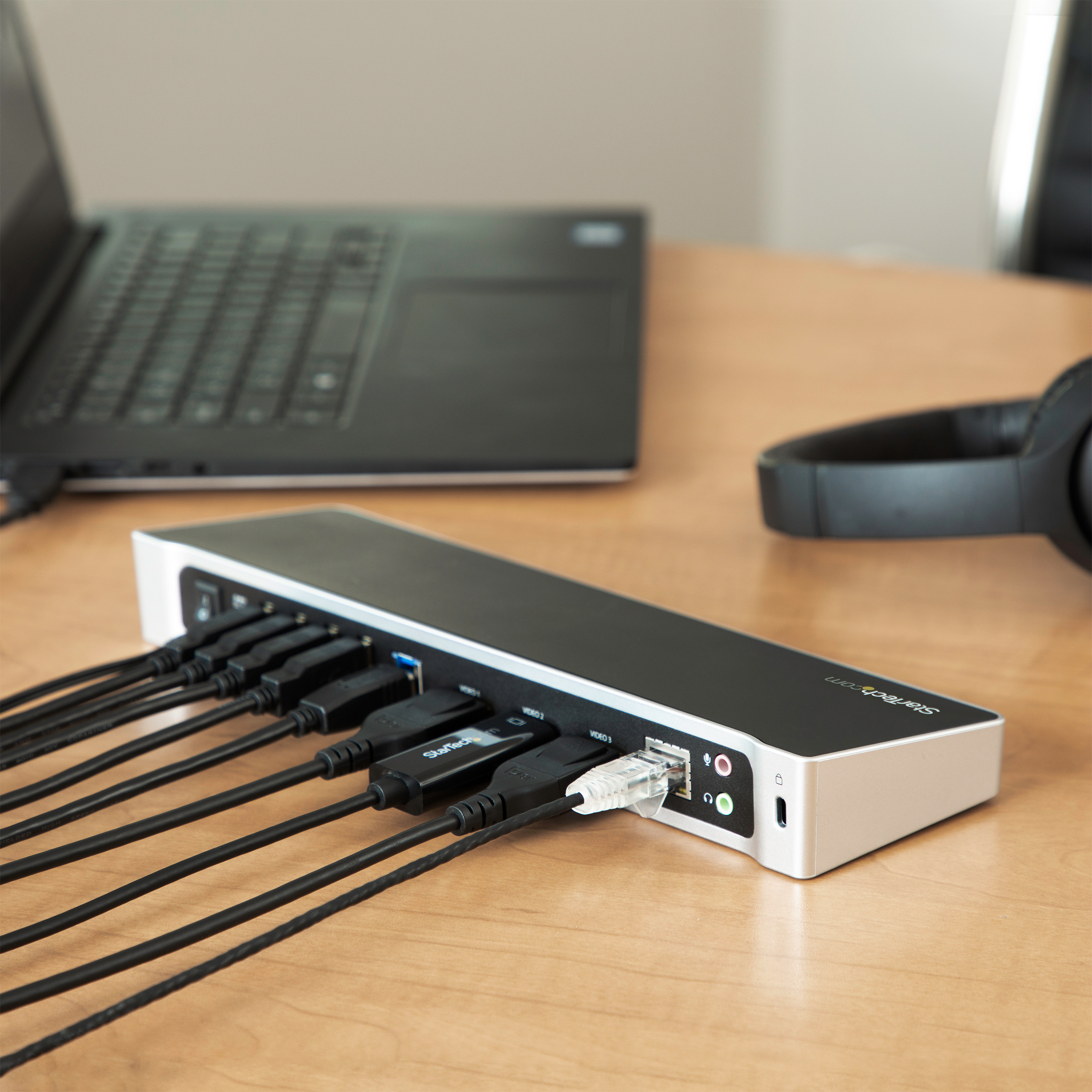 Una docking station nera con vari cavi collegati e un laptop sullo sfondo.