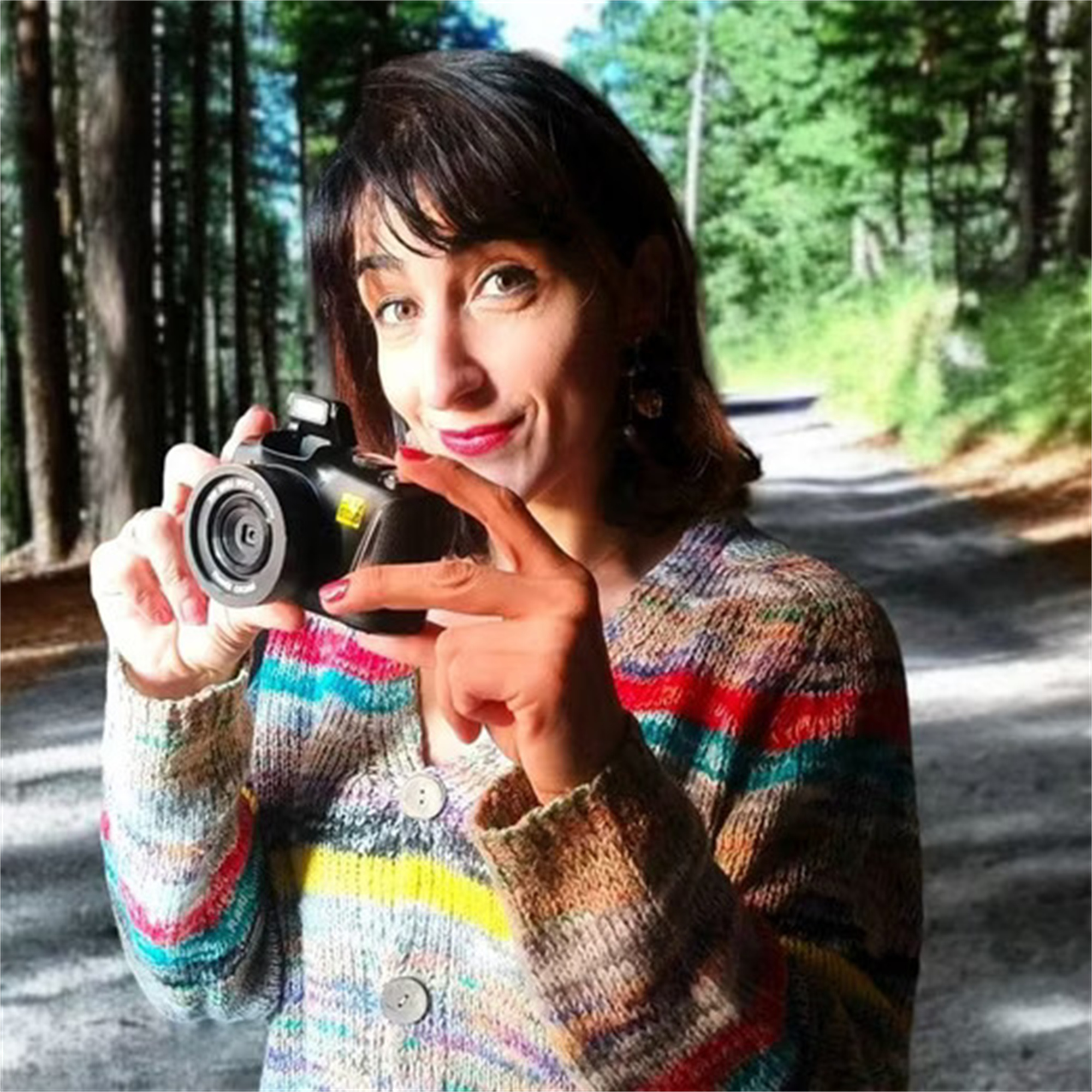 Frau mit Kamera in Waldumgebung, lächelnd, hält Kamera, Bäume im Hintergrund.