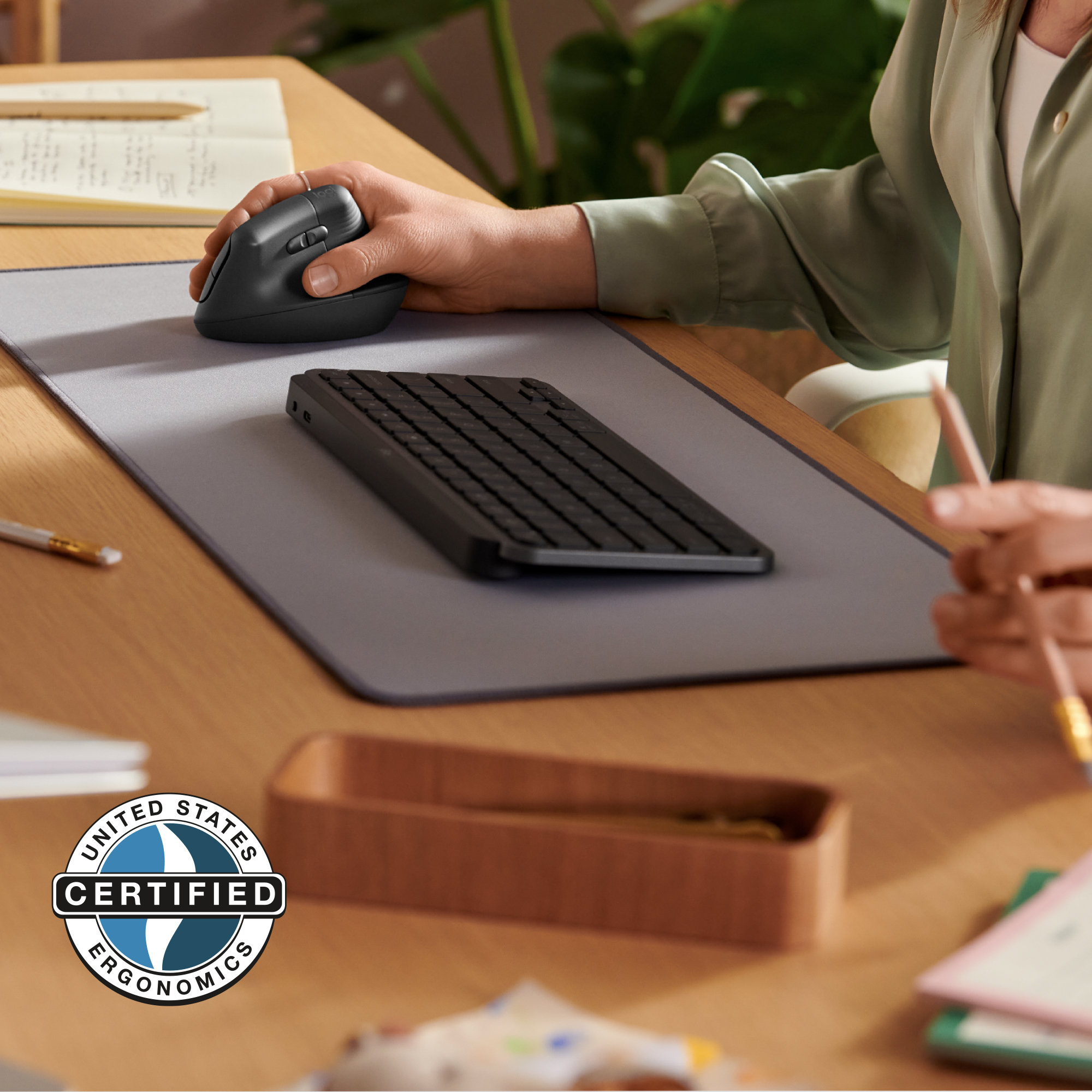 Osoba używająca czarnej myszy i klawiatury na drewnianym biurku. Widoczna etykieta Certified Ergonomics.