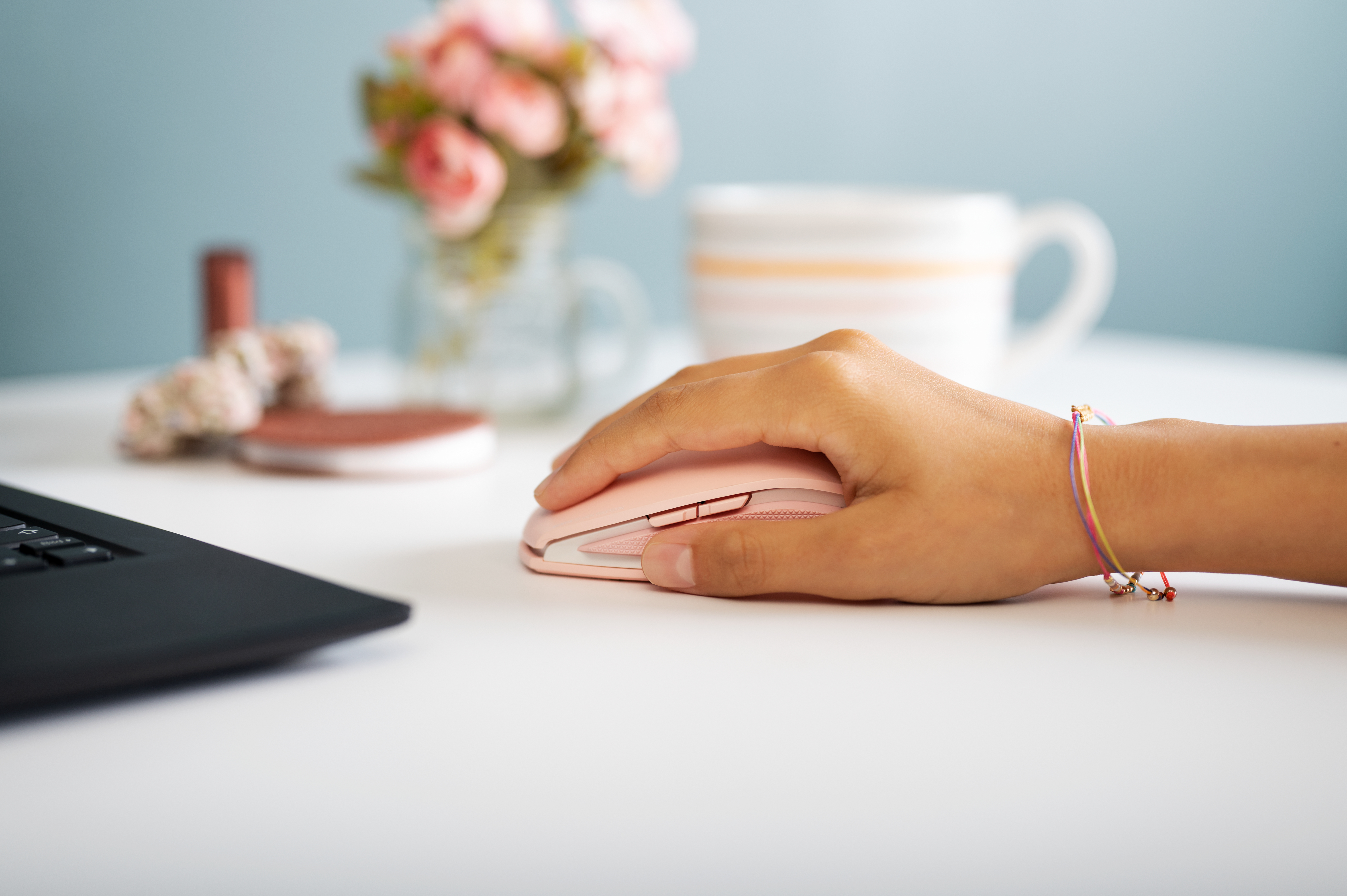 A person's hand using a pink computer mouse on a white desk. A cup and monitor are in the background.