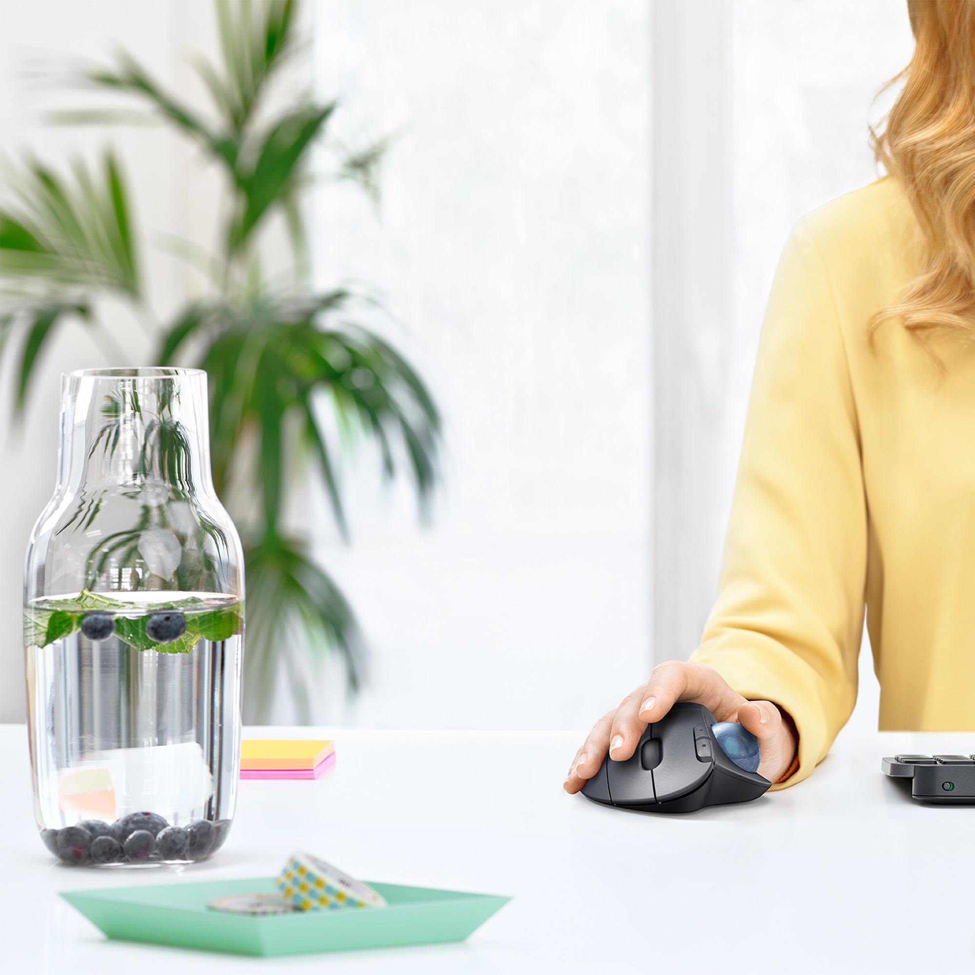 Kobieta używa myszy komputerowej na białym biurku. Szklanka wody i roślina w tle.