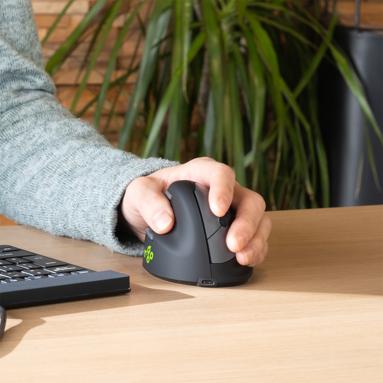 Person benutzt eine vertikale Maus auf einem Holzschreibtisch. Eine Tastatur ist im Vordergrund.