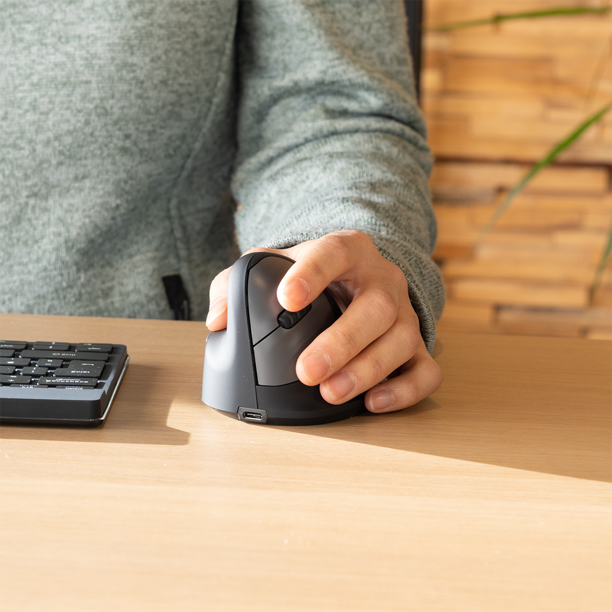 Die Hand einer Person benutzt eine vertikale ergonomische Maus auf einem Holzschreibtisch neben einer Tastatur.