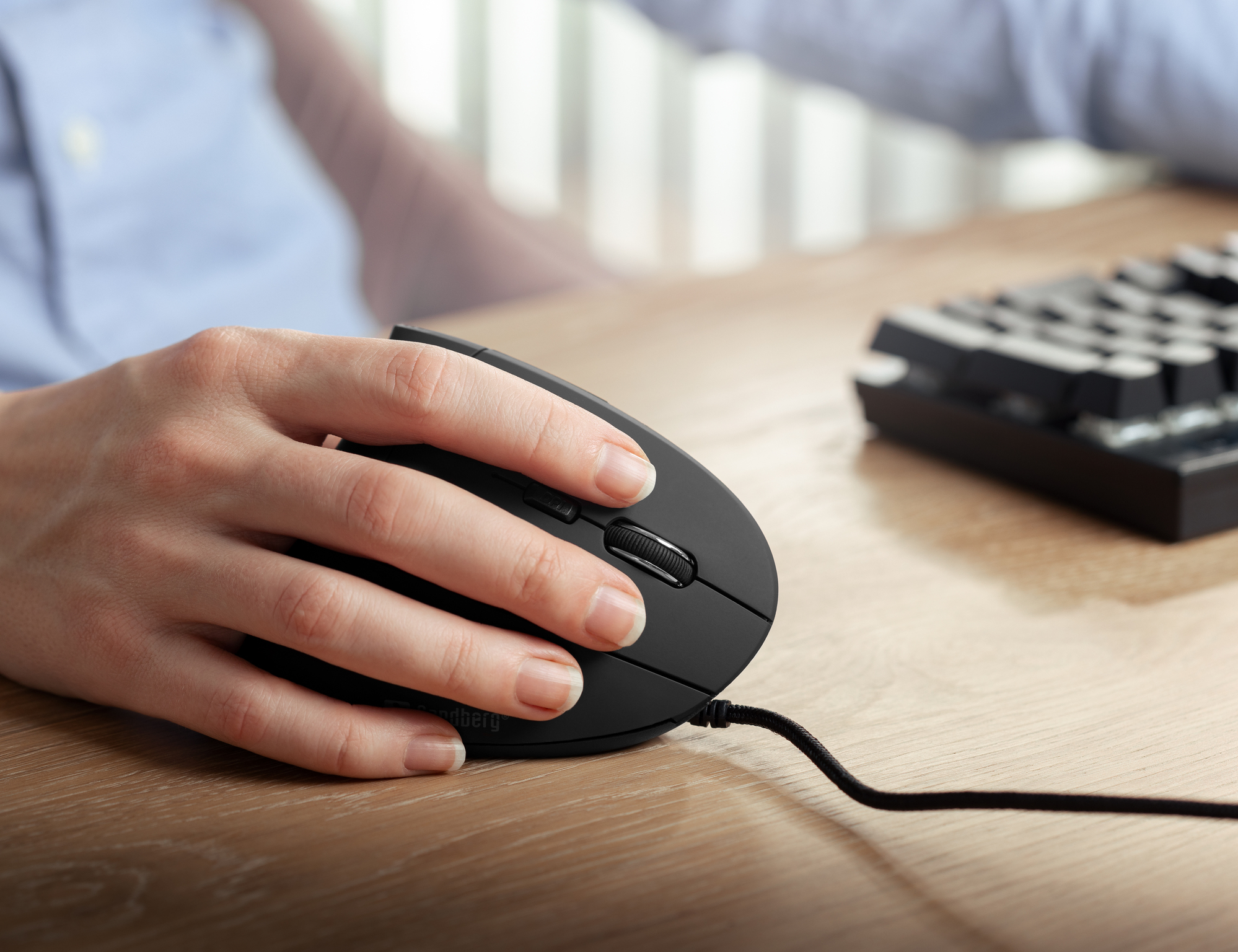 Ręka używająca czarnej myszy. Mysz znajduje się na drewnianej powierzchni, w tle klawiatura.