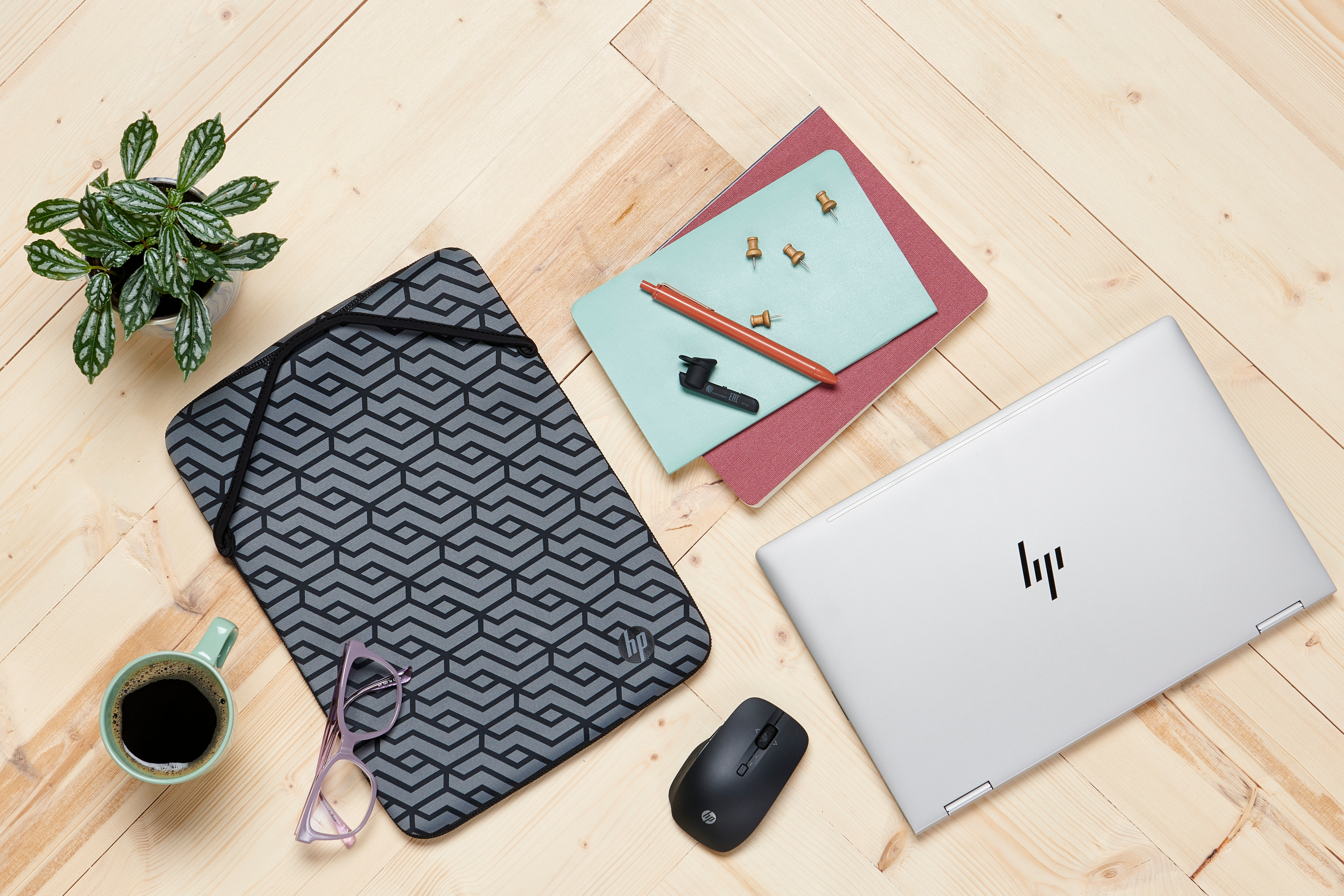 Un laptop, un mouse, una custodia, un taccuino, occhiali e una tazza su un tavolo di legno con una pianta.