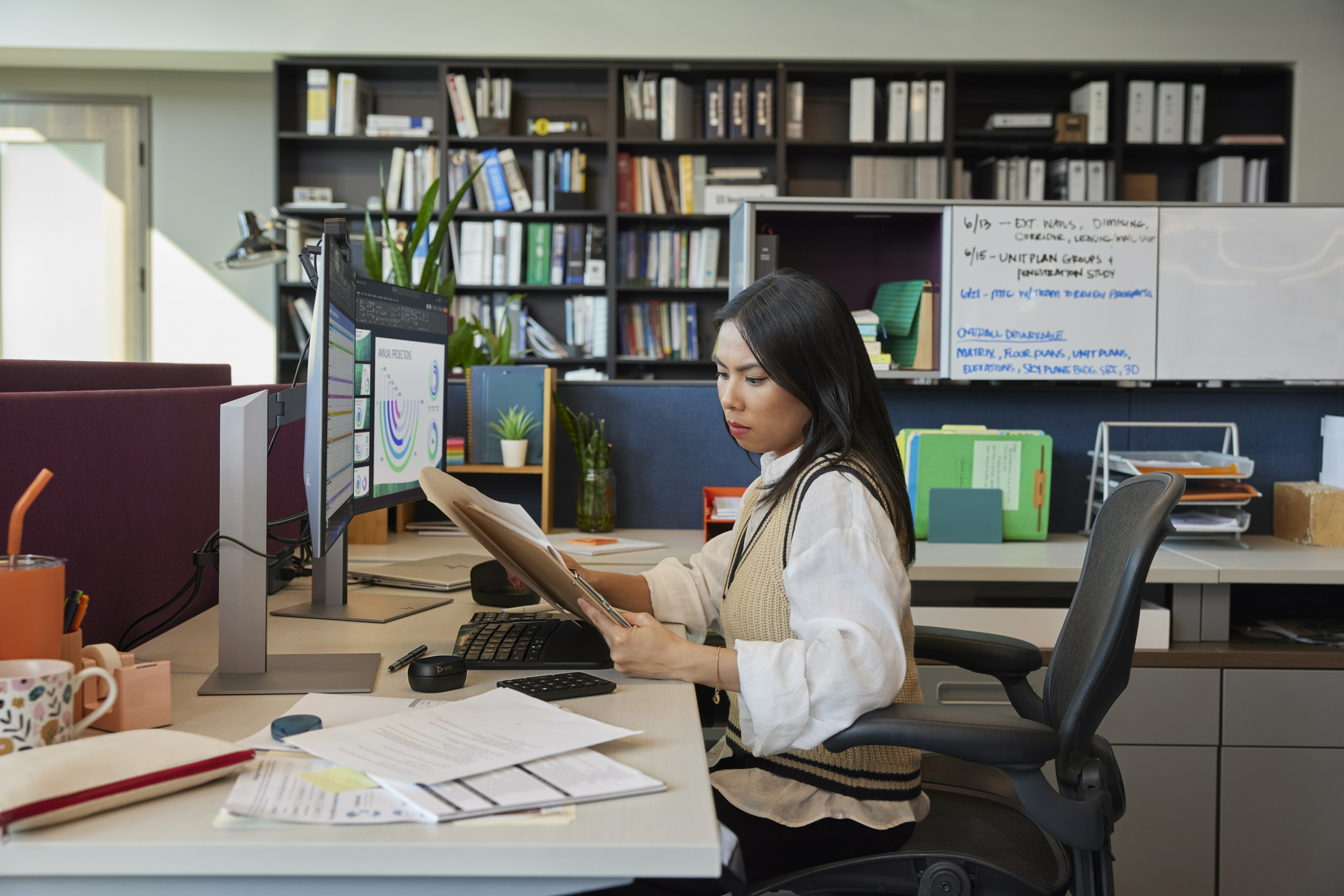 Kobieta w biurze przegląda papiery, z biurkiem, komputerem i regałem z książkami w tle.