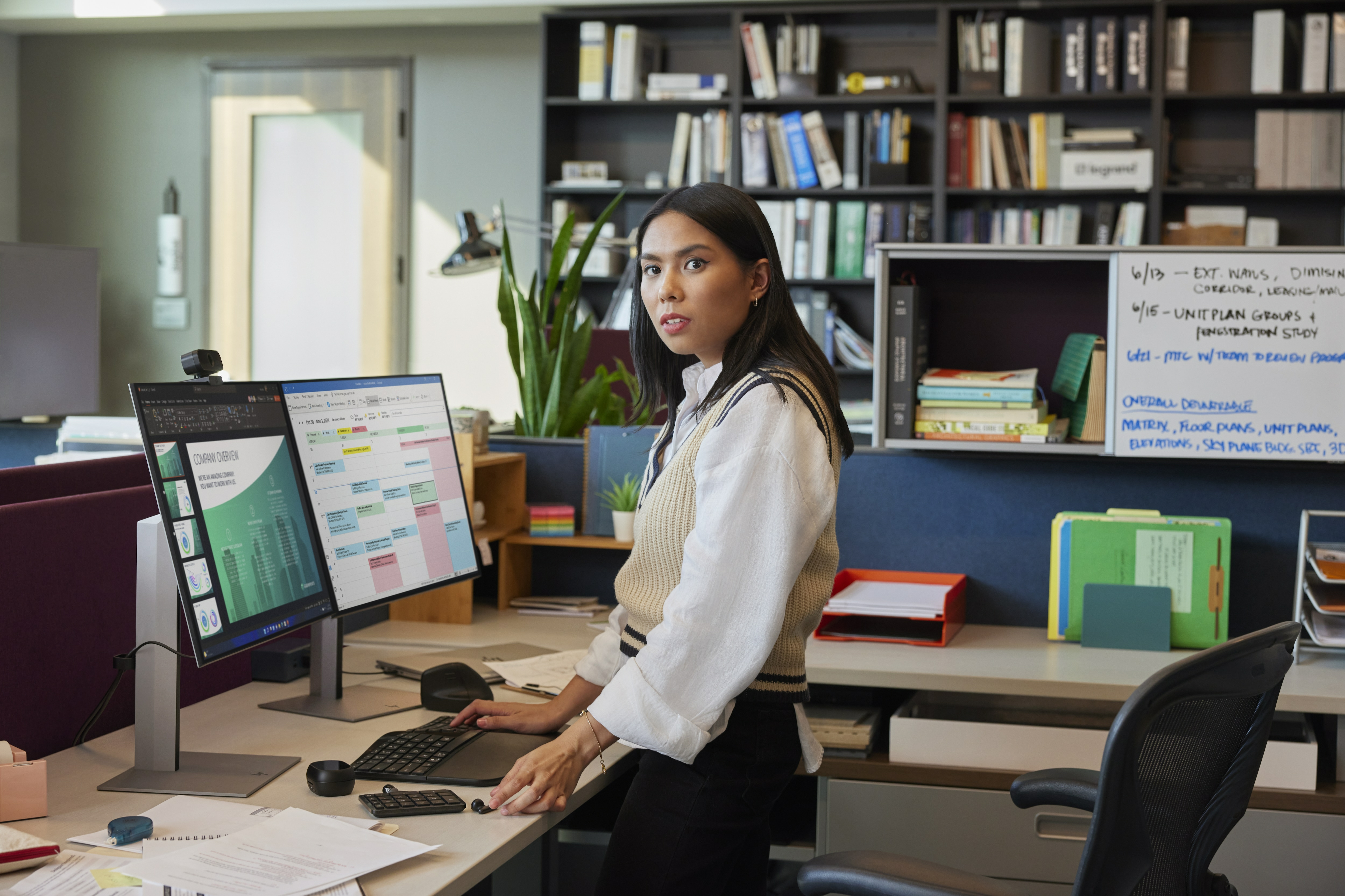 Kobieta w biurze patrzy w kamerę, z dwoma monitorami i półką z książkami w tle.