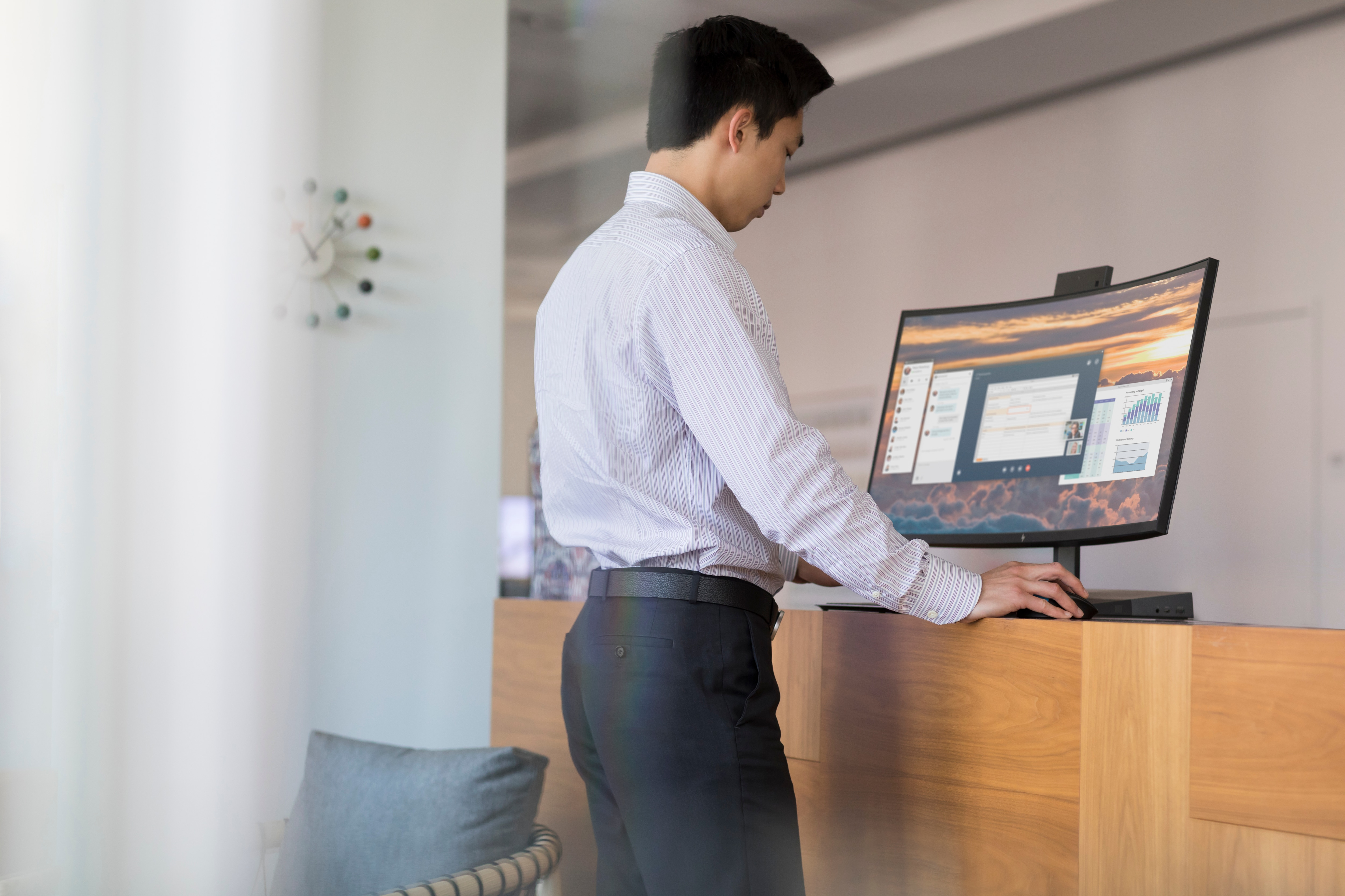 Mężczyzna używa komputera na biurku. Monitor wyświetla dane. Zegar i roślina po lewej stronie.