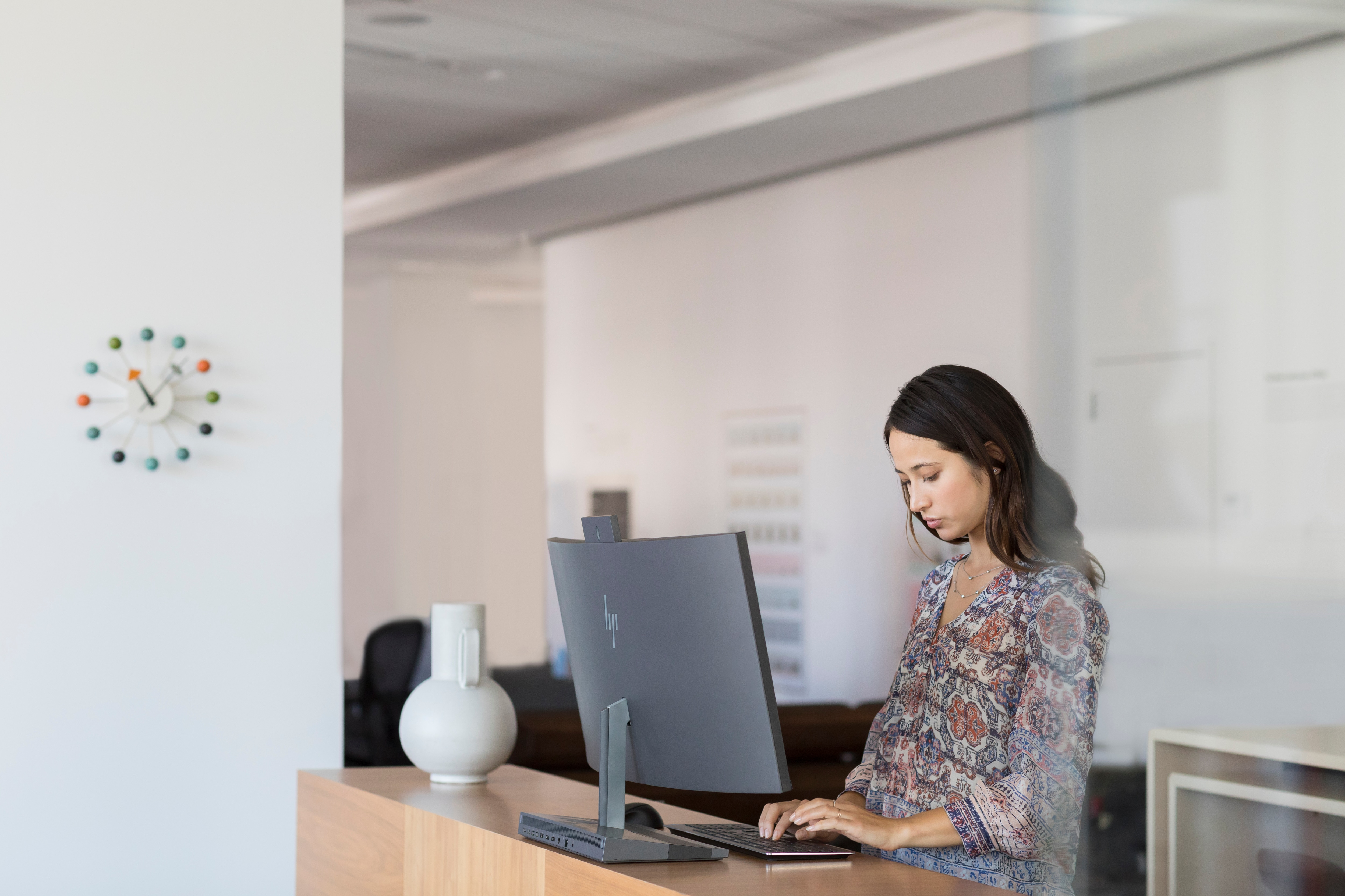 Kobieta pisze na komputerze przy biurku. Zegar i wazon są na ścianie.
