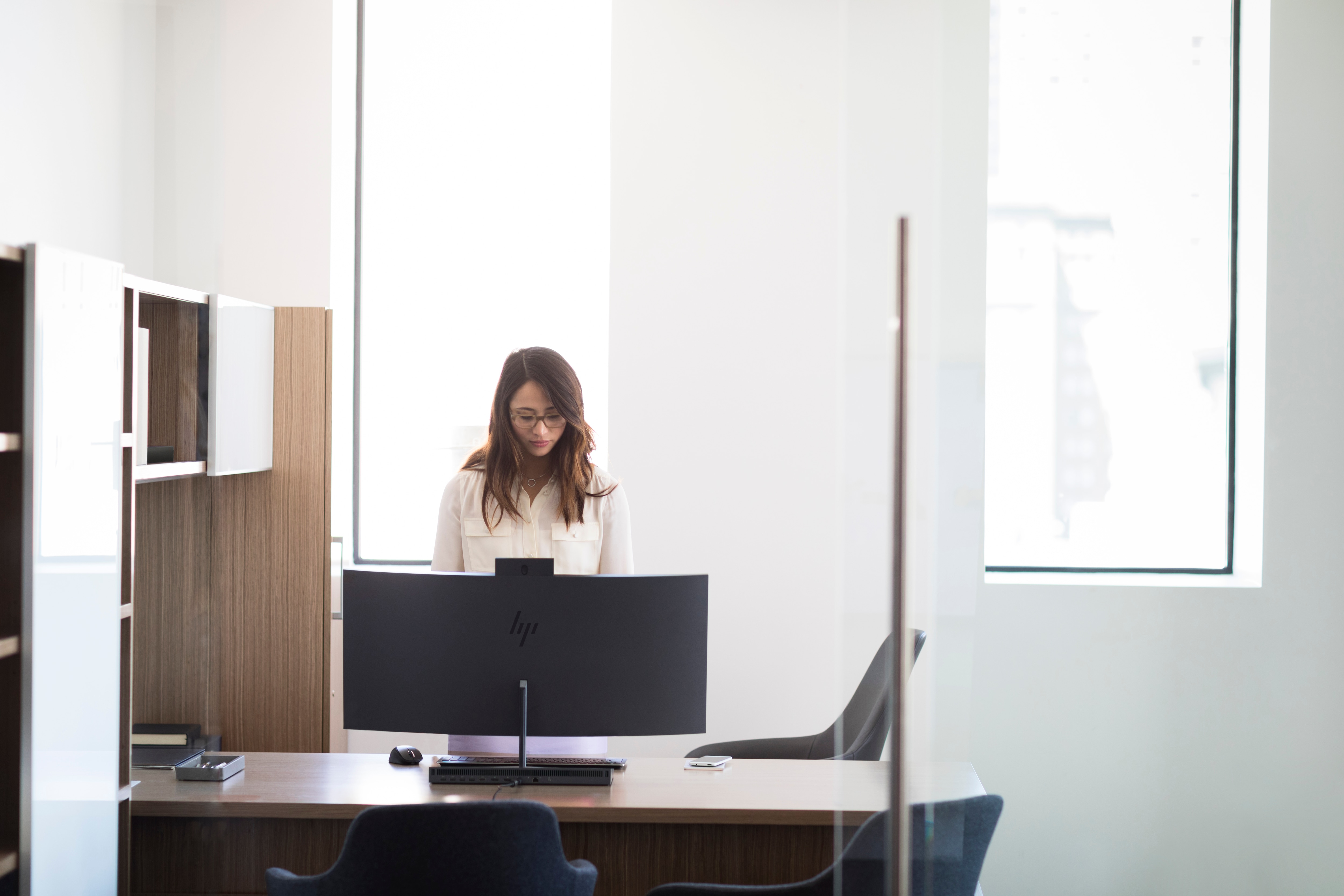 Kobieta w okularach pracuje przy biurku z komputerem, patrząc w dół, w otoczeniu biurowym.