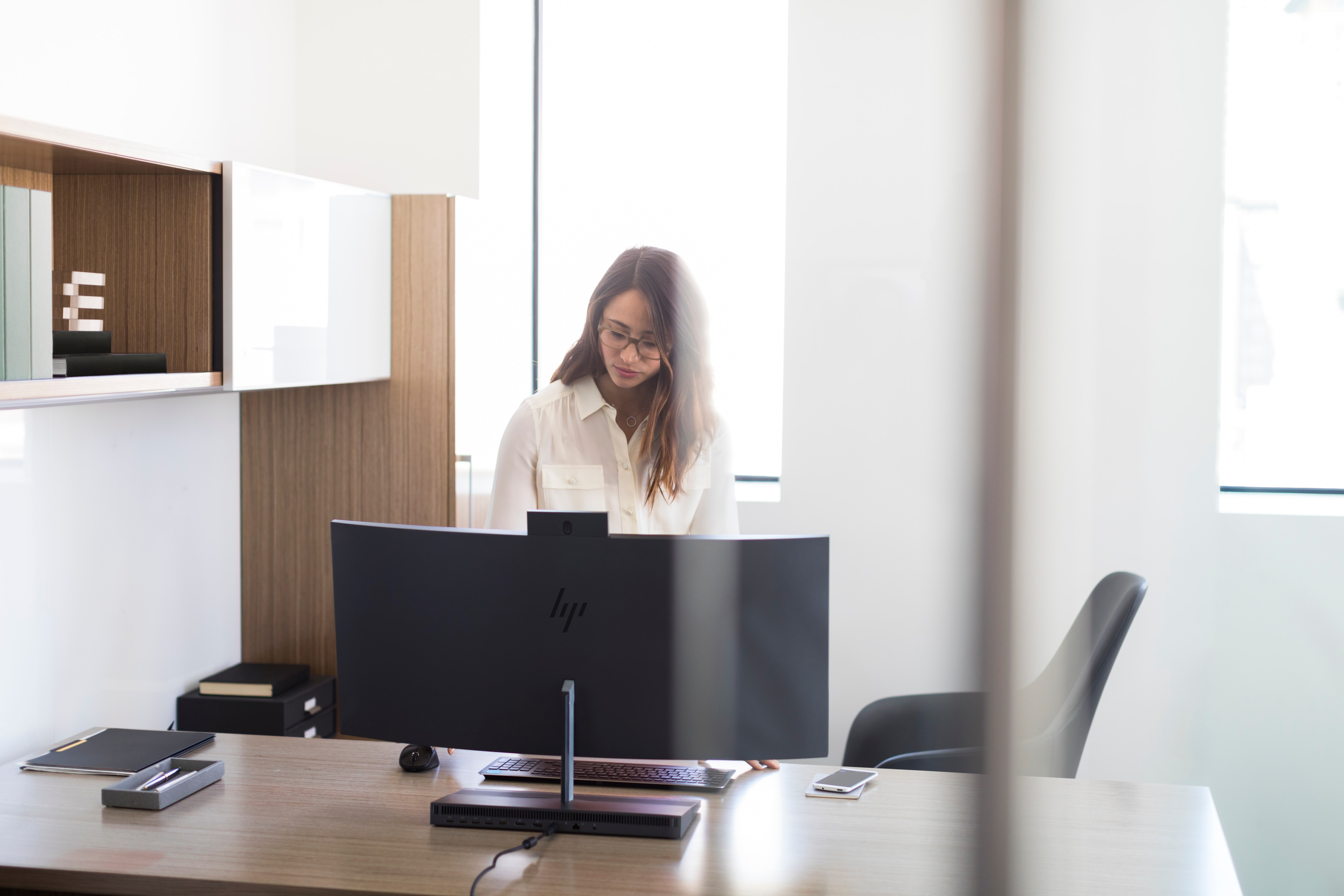 Kobieta w okularach pracująca przy biurku z dużym zakrzywionym monitorem.