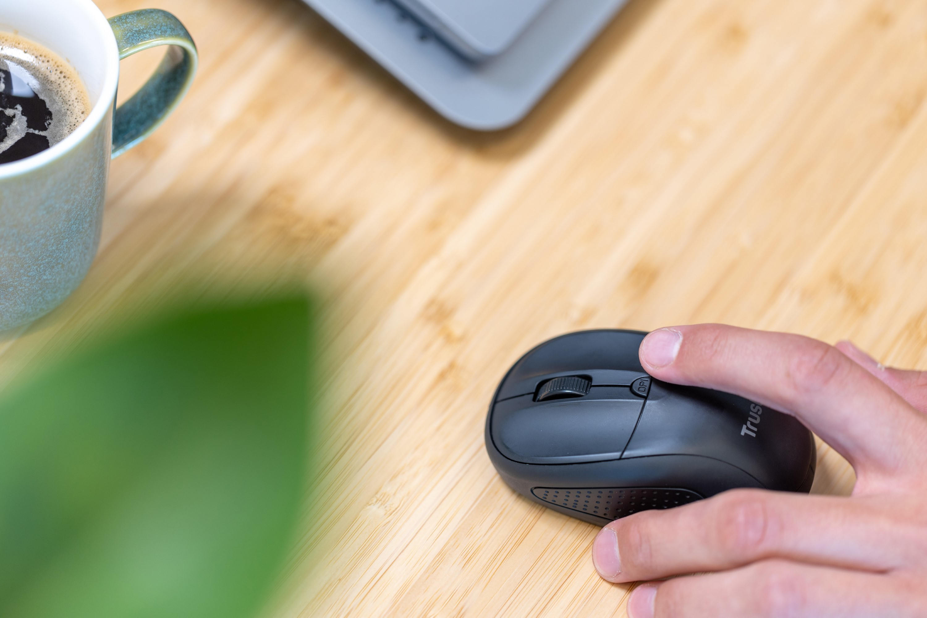 Main utilisant une souris noire sur une table en bois près d'une tasse et plante verte.
