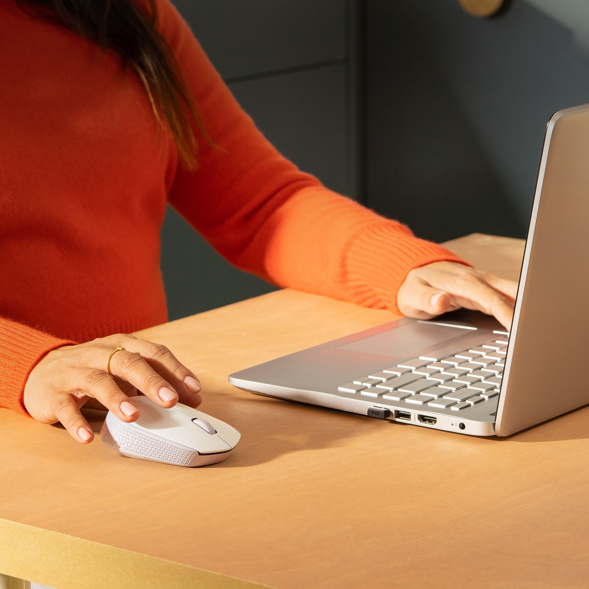 Osoba w pomarańczowym swetrze używa laptopa i białej myszy na drewnianym stole.