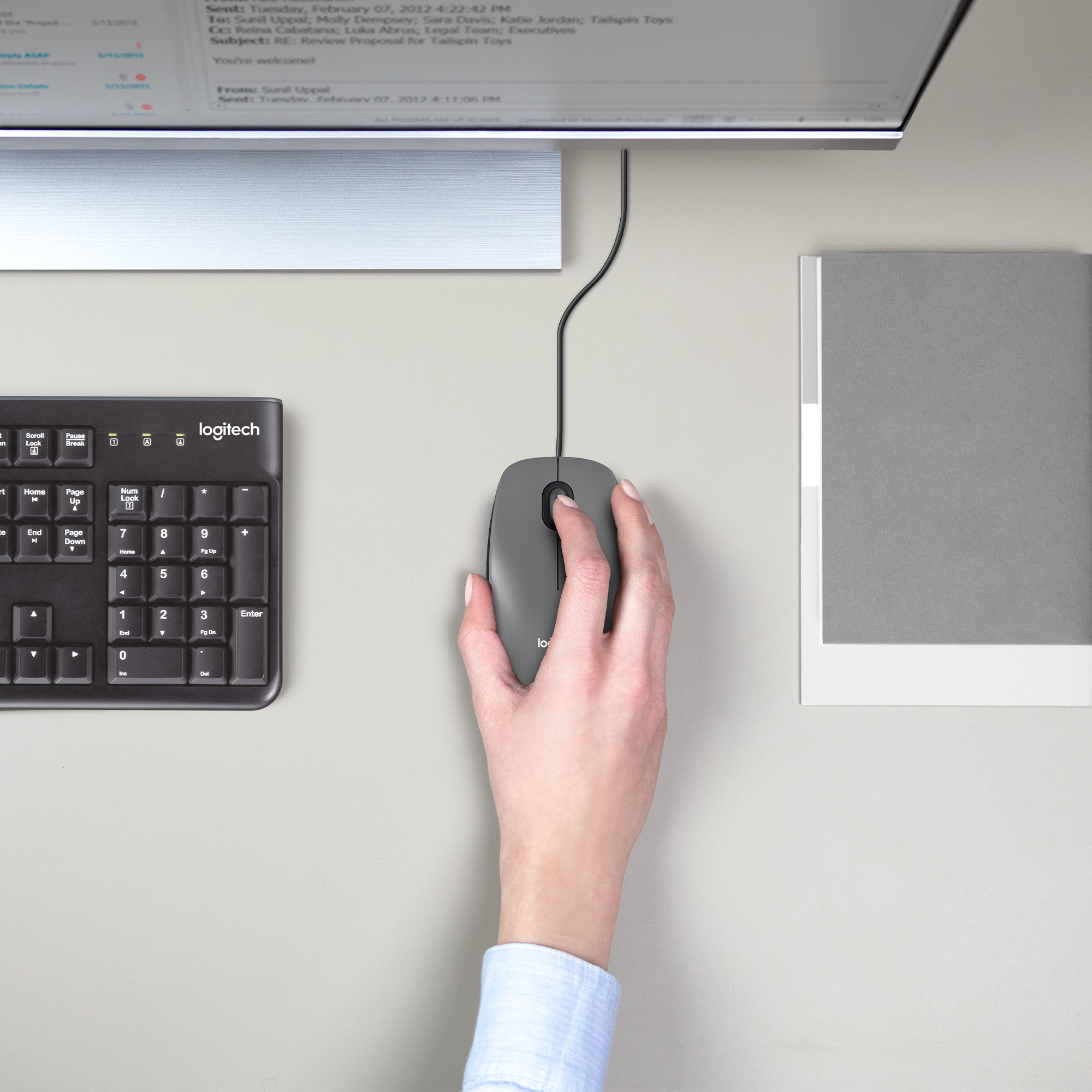 Eine Hand benutzt eine graue Maus auf einem Schreibtisch mit Tastatur und Monitor. Die Maus ist kabelgebunden.