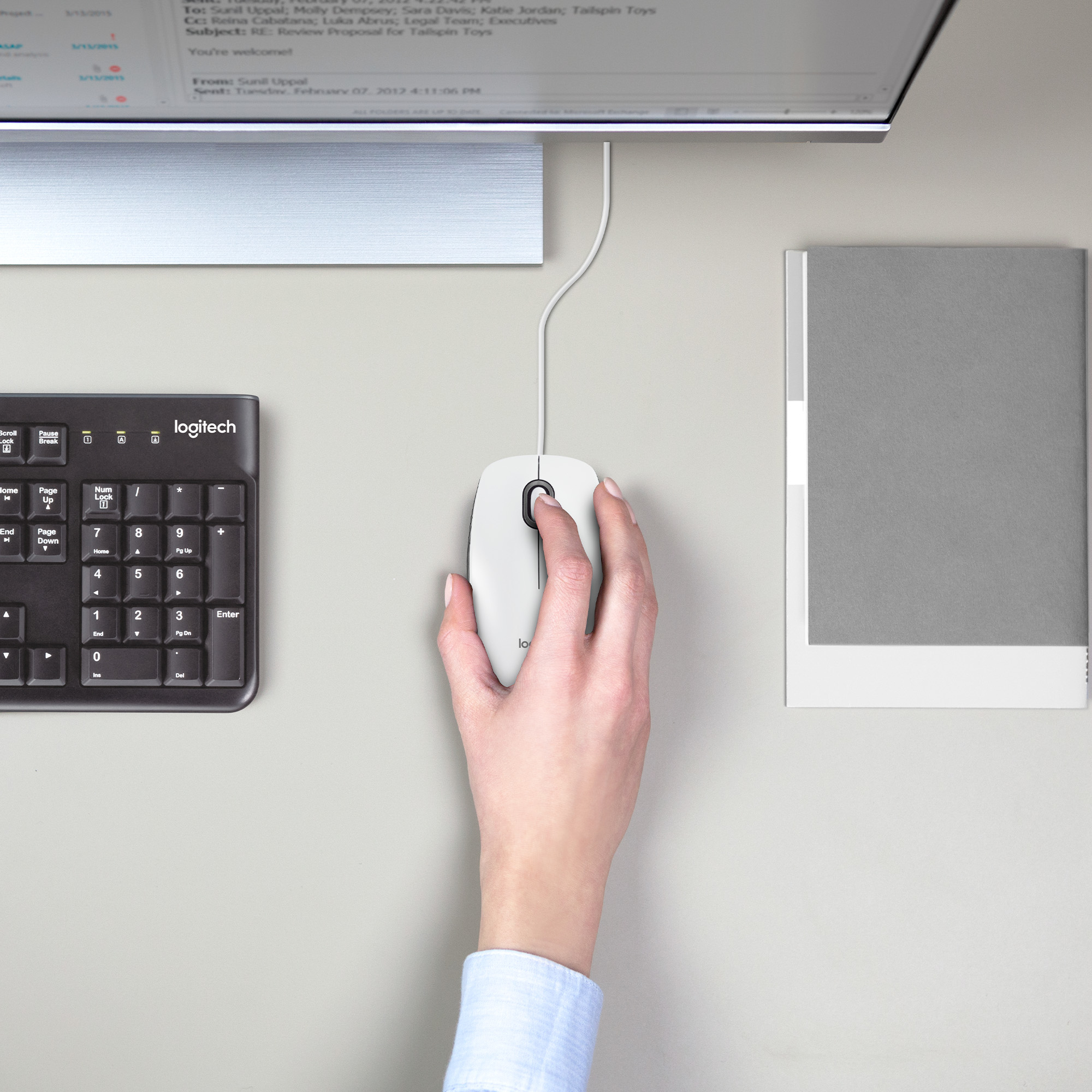 Ręka używająca białej myszy Logitech. Klawiatura i monitor w tle.