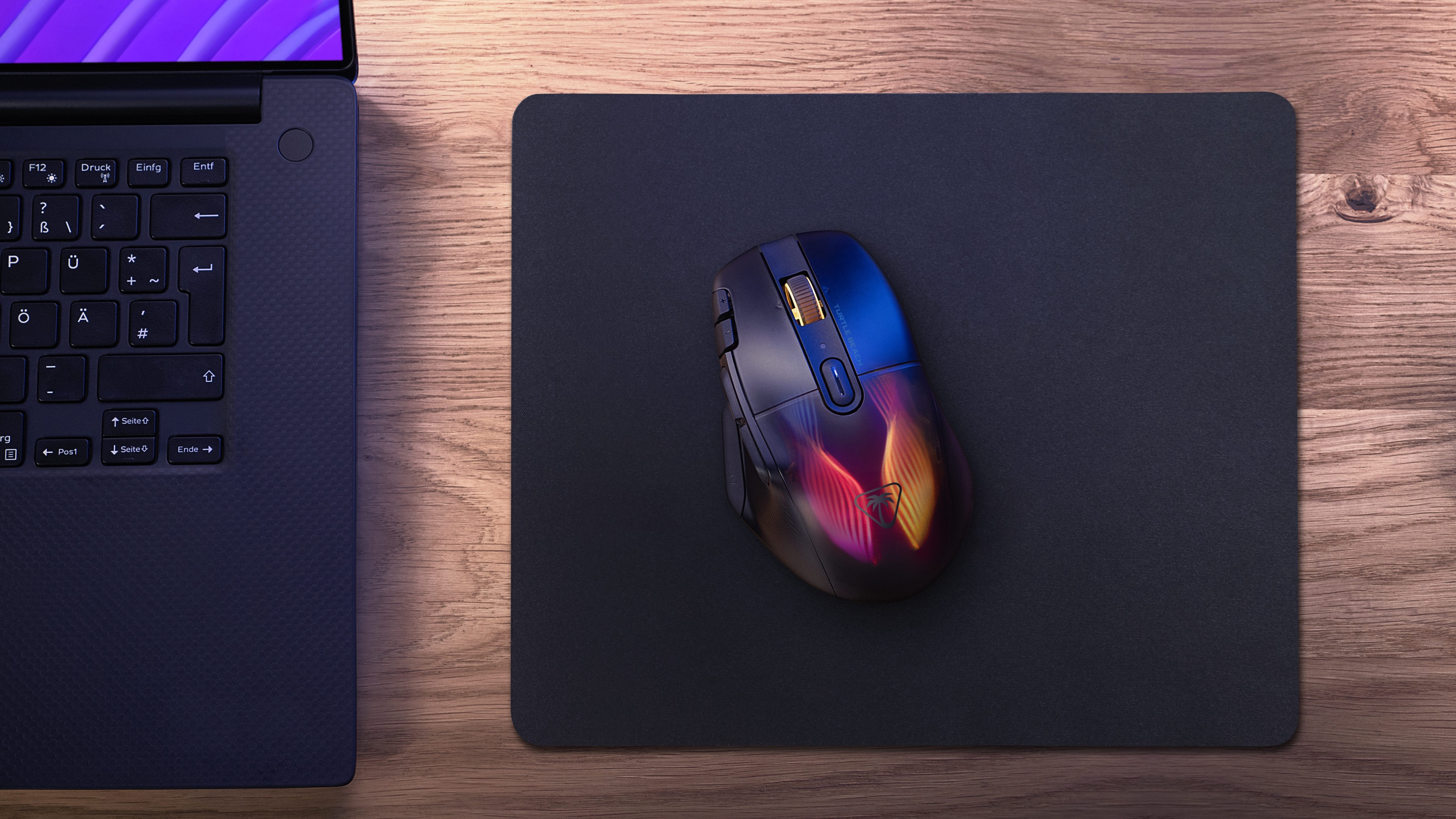 Een gamingmuis op een muismat op een houten bureau. Een laptop staat links. Warme verlichting.