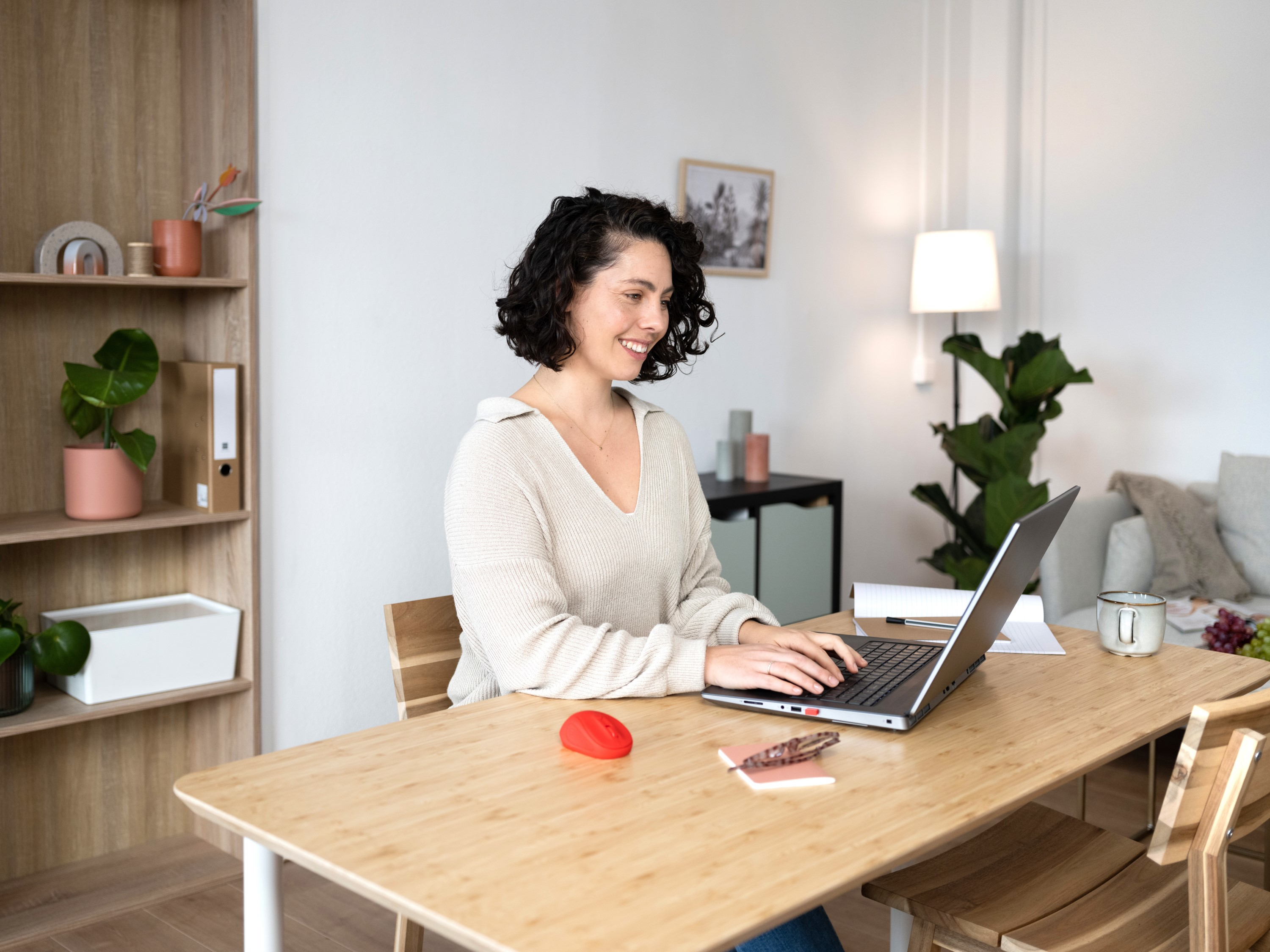 Donna che lavora su un laptop a un tavolo di legno con mouse rosso, sorridente e sfondo stanza.